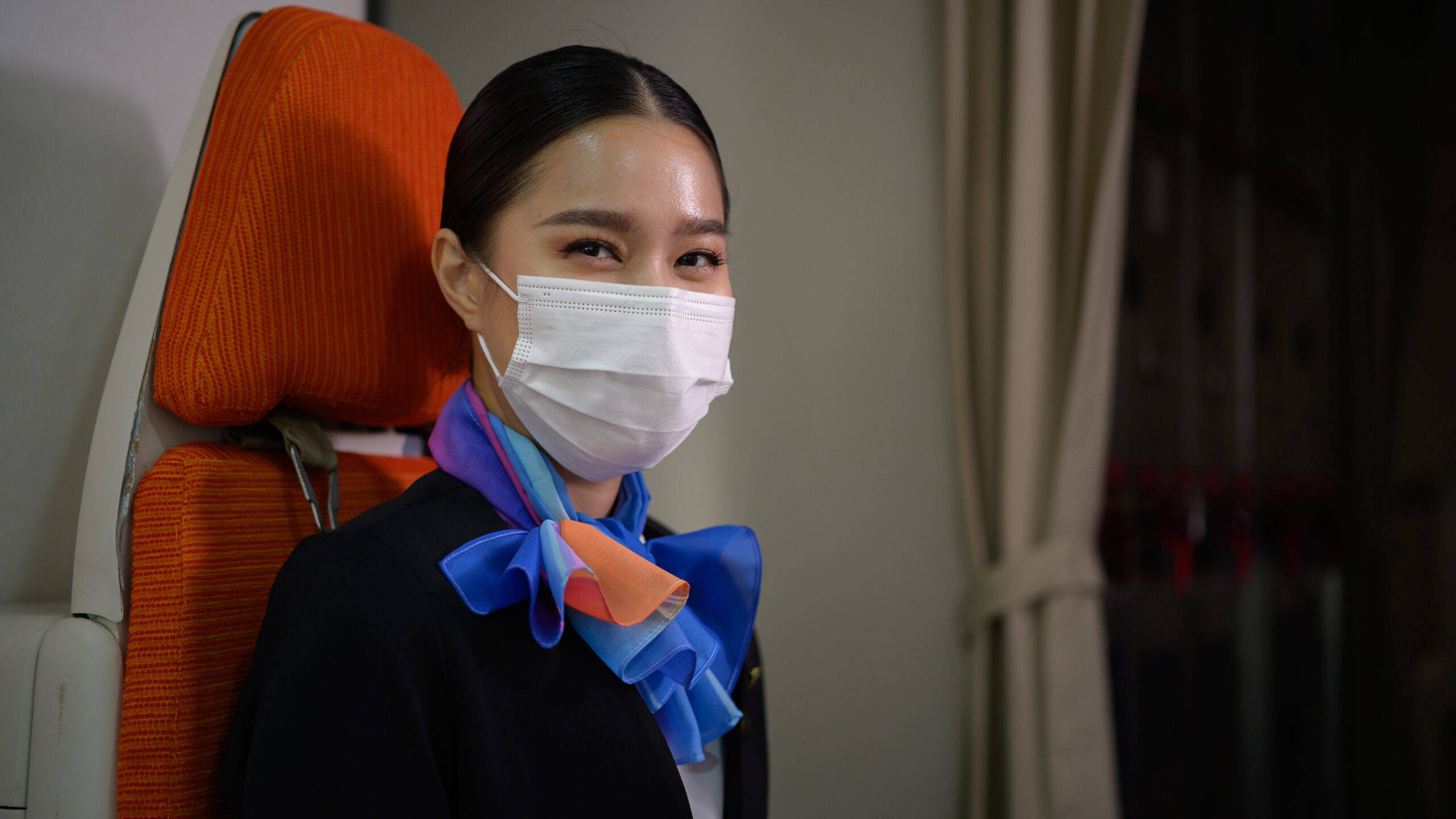 Airport 桌布 壁紙 — 一位戴著白色口罩和彩色圍巾的空中服務員，平靜地坐在橙色座椅的飛機座位上。