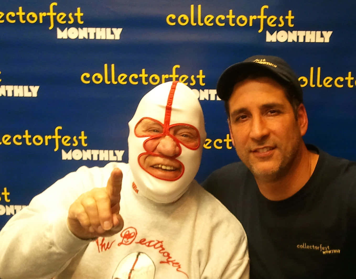 WWE 桌布 壁紙 — 一位戴著白色和紅色面具的蒙面摔角手，在一個微笑戴黑色帽子的男子旁邊，指向前方，攝於Collectorfest Monthly活動。