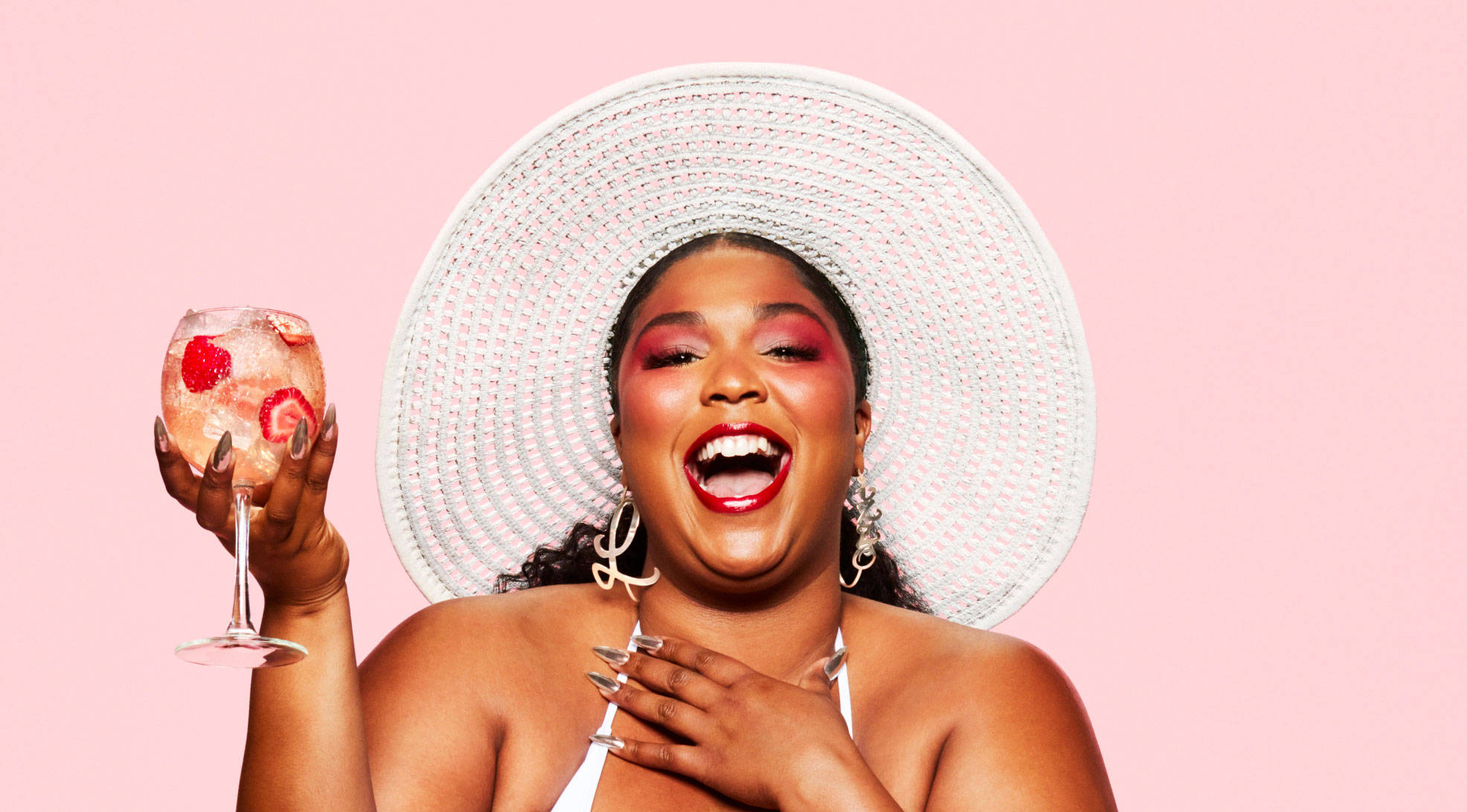 Lizzo 桌布 壁紙 — 一位戴著白色太陽帽的快樂女性，在柔和的粉紅色背景前，手持一杯粉紅色雞尾酒笑著。