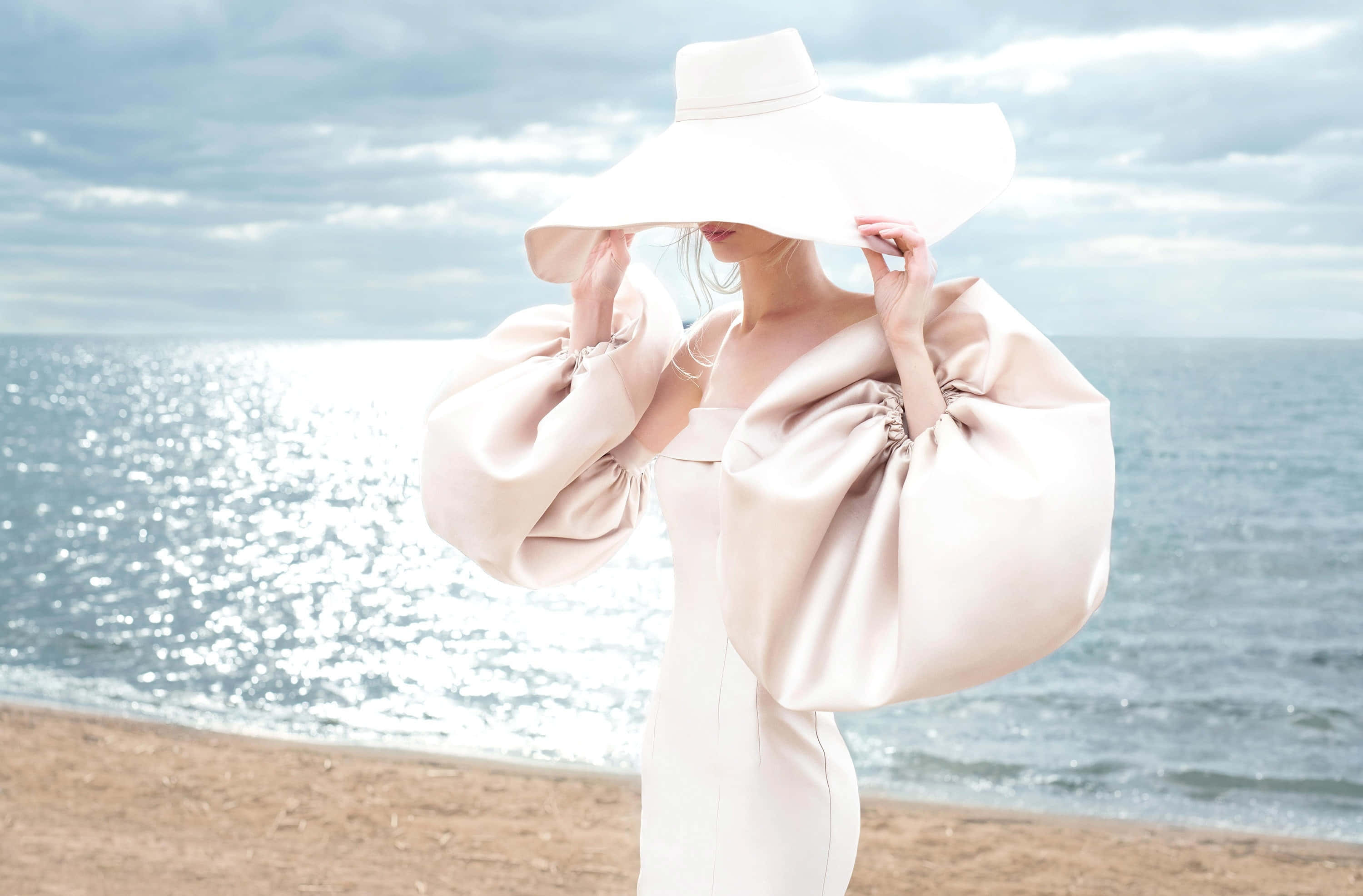 Outdoor Activities 桌布 壁紙 — 一位戴著白色寬邊帽、身穿飄逸米色長袍的女性，優雅地站在陽光普照的海灘上，身後是閃閃發光的海浪。