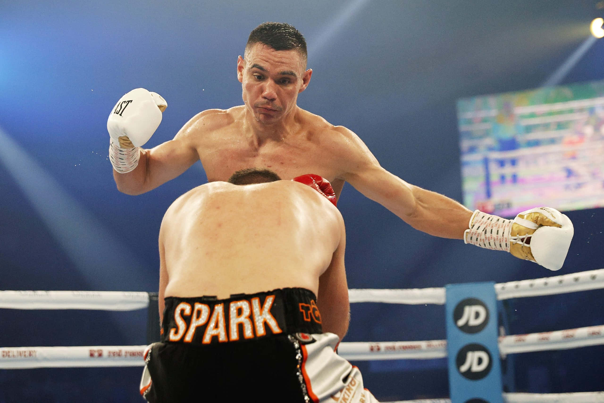 Tim Tszyu 桌布 壁紙 — 一位戴著白色手套的拳擊手在燈光明亮的擂台上，向對手的背部猛烈出拳。
