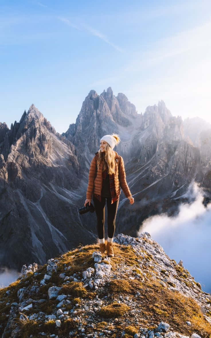 Winter Hiking 桌布 壁紙 — 一位戴著白色毛帽和棕色外套的女性站在岩石山頂上微笑，雲朵在她下方飄過，襯著清澈的藍天。