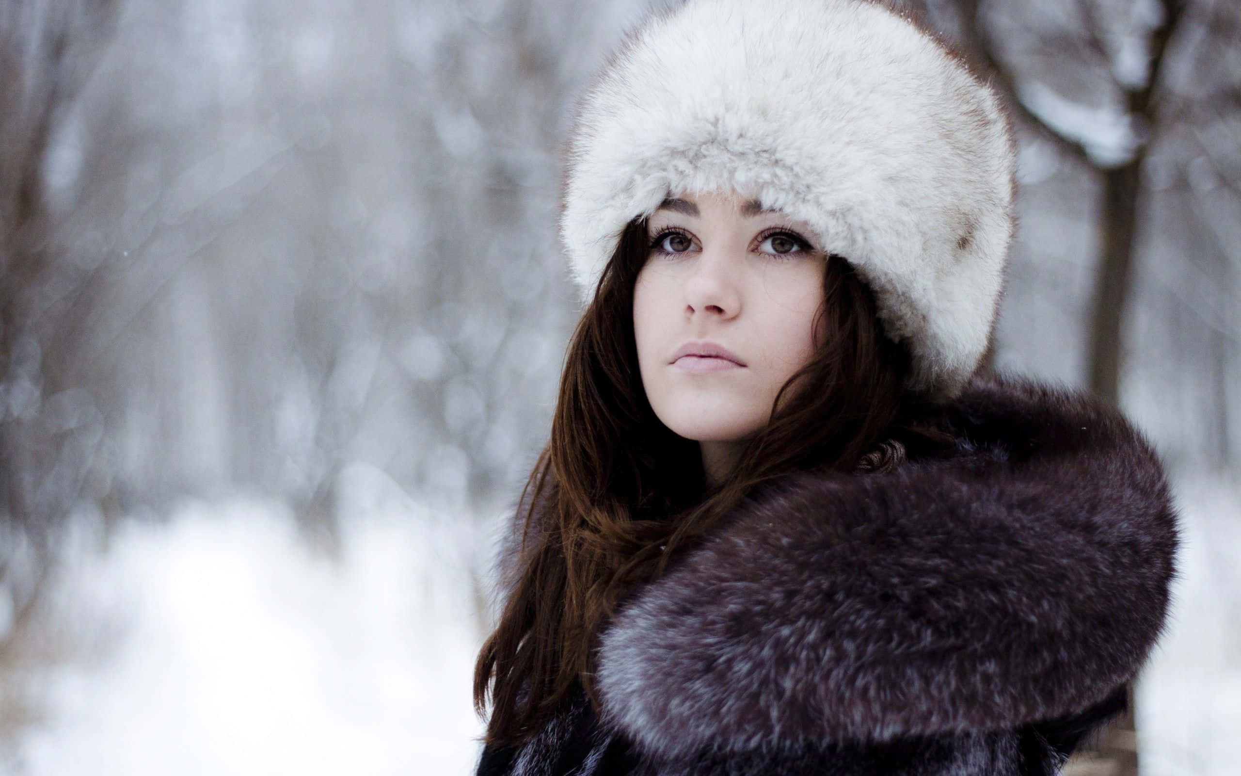 Winter Fashion 桌布 壁紙 — 一位戴著白色毛帽和深色毛大衣的女性，透過身後樹木模糊的雪林若有所思地凝視。