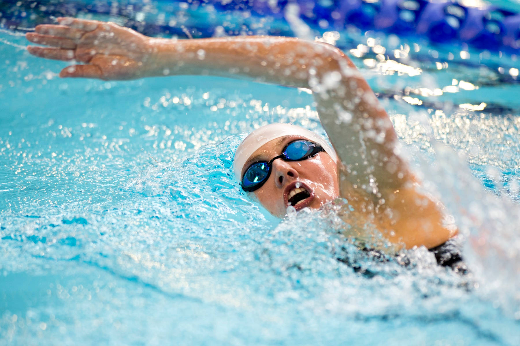 Swimming 桌布 壁紙 — 一位戴著白色泳帽和藍色泳鏡的泳者，在明亮的藍色泳池水中強力划水，濺起的水滴捕捉到陽光。