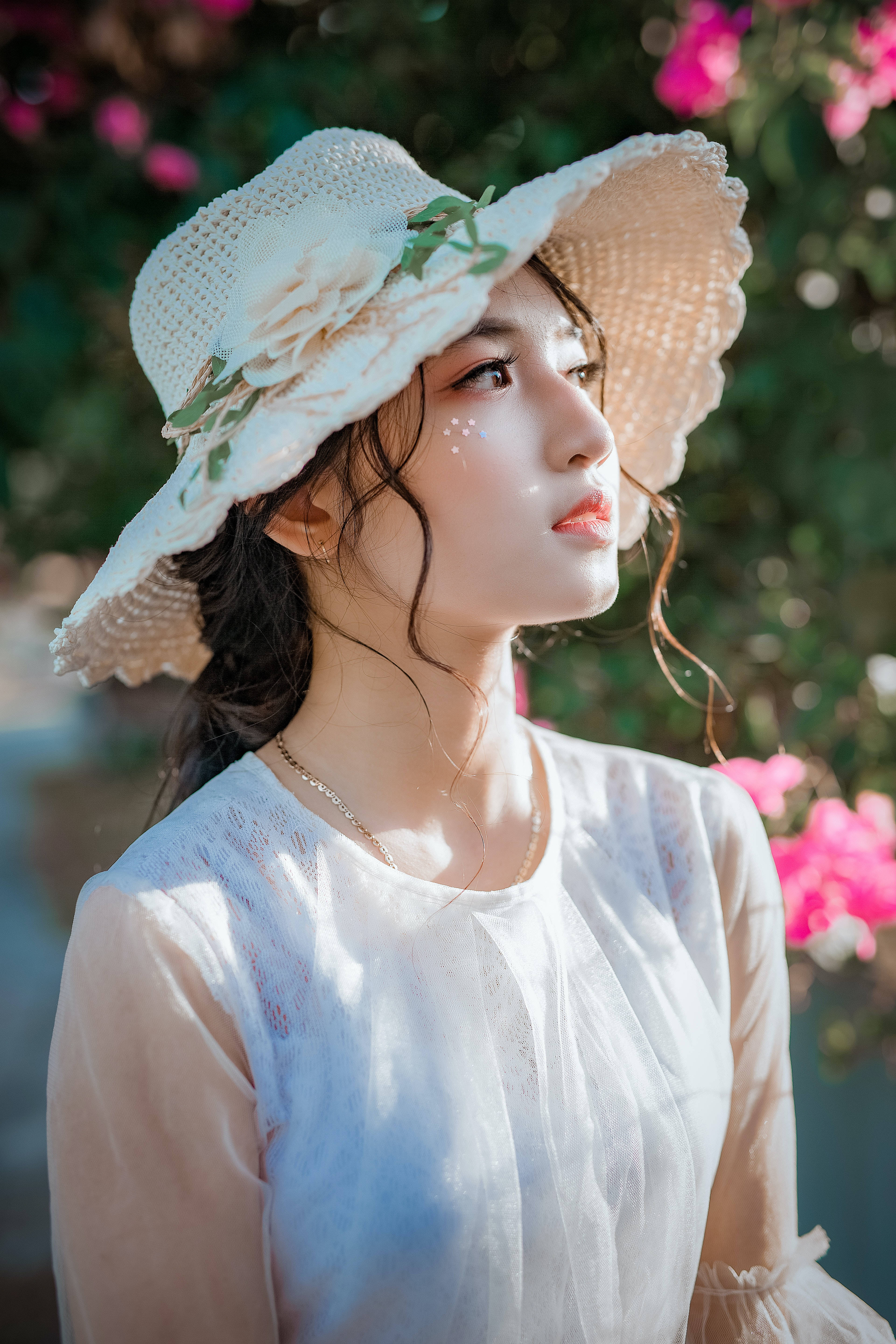 美女 桌布 壁紙 — 一位戴著白色蕾絲帽和透視襯衫的年輕女子在柔和的粉紅色花朵和斑駁的陽光中向上凝視。