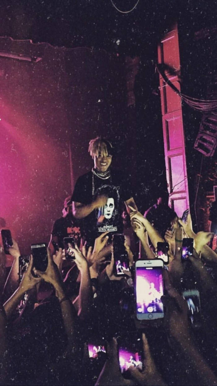 xxxtentacion 美學 桌布 壁紙 — 一位戴著白色面具的表演者在粉紅色舞台燈光下，在一群高舉發光手機的粉絲上方咧嘴而笑。