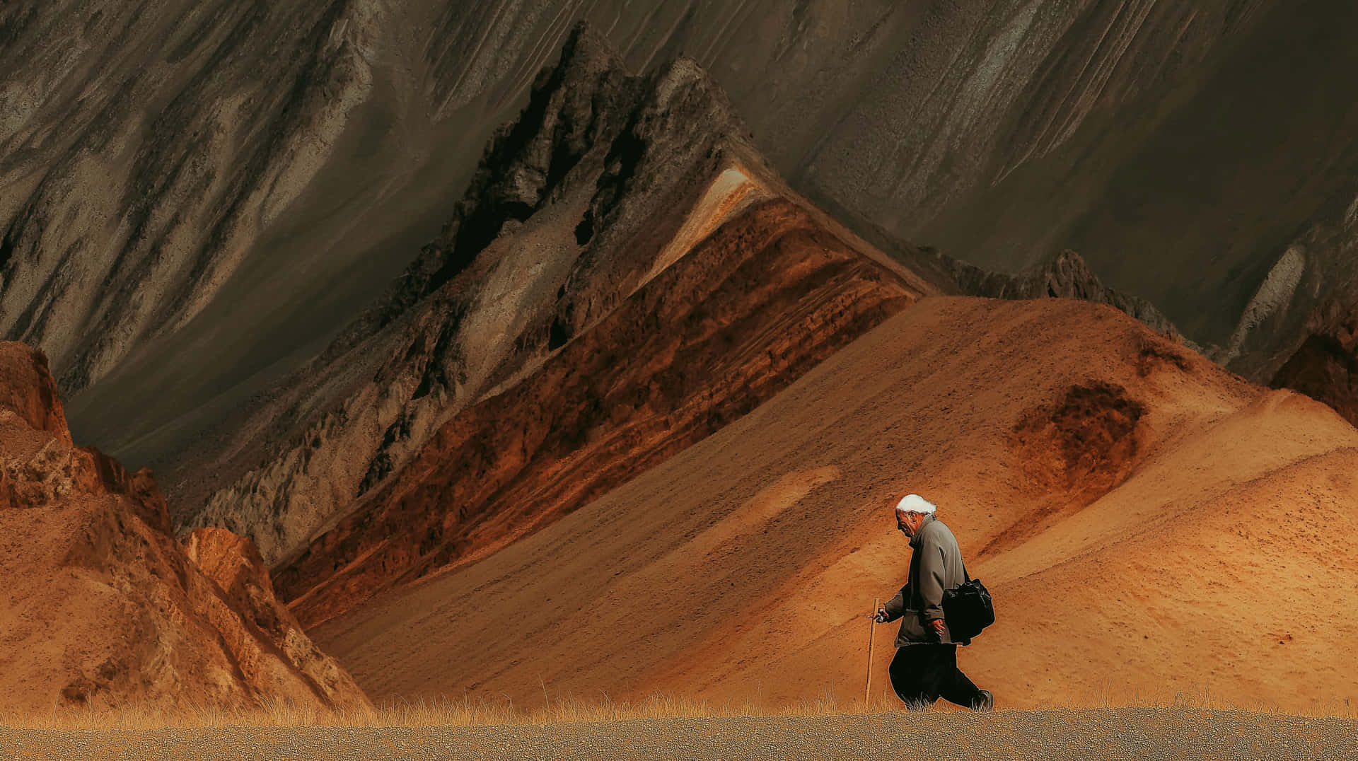Solitary Figure Journeying Through Mountainous Vistas 壁紙 — 一位戴著白色頭巾、拄著拐杖的長者獨自走過廣闊乾旱的景觀，層層疊疊的橙色和灰色山脈在暗淡的天空下。