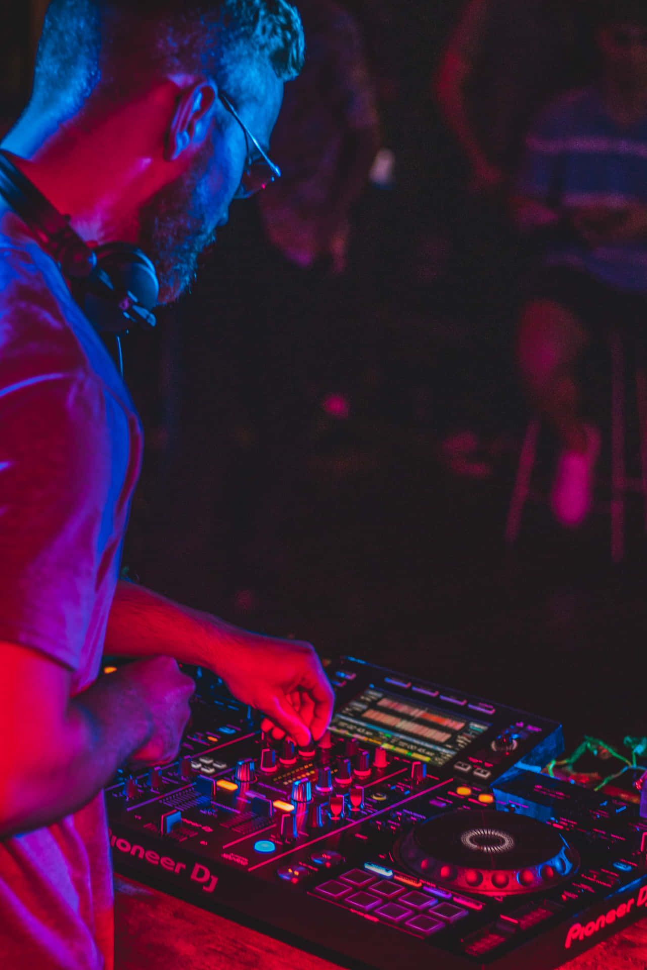 DJ 桌布 壁紙 — 一位戴著眼鏡和耳機的DJ在充滿活力的紅色和藍色俱樂部燈光下，使用Pioneer DJ控制器混音音樂。