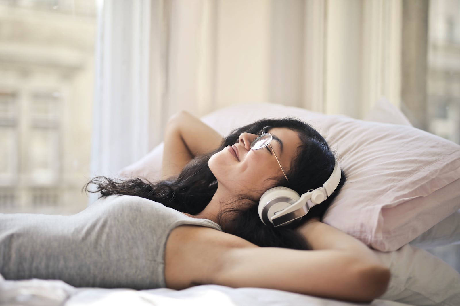 Relaxing 桌布 壁紙 — 一位戴著眼鏡和耳機的女性在明亮的窗邊，在柔軟枕頭的床上平靜地休息。