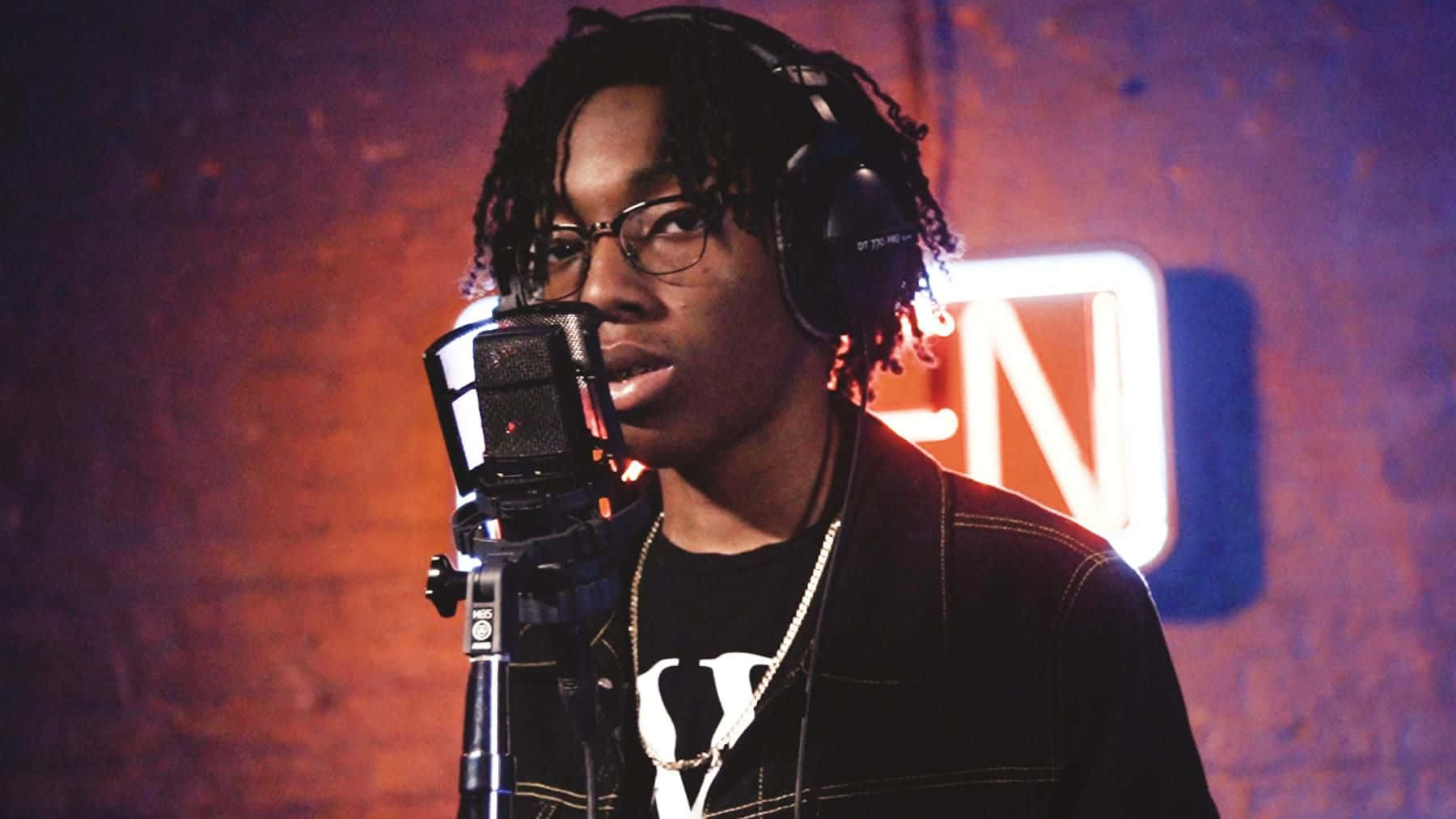 Rappers 桌布 壁紙 — 一位戴著眼鏡和耳機的年輕饒舌歌手在昏暗的錄音室裡，在溫暖的霓虹燈下對著麥克風唱歌。