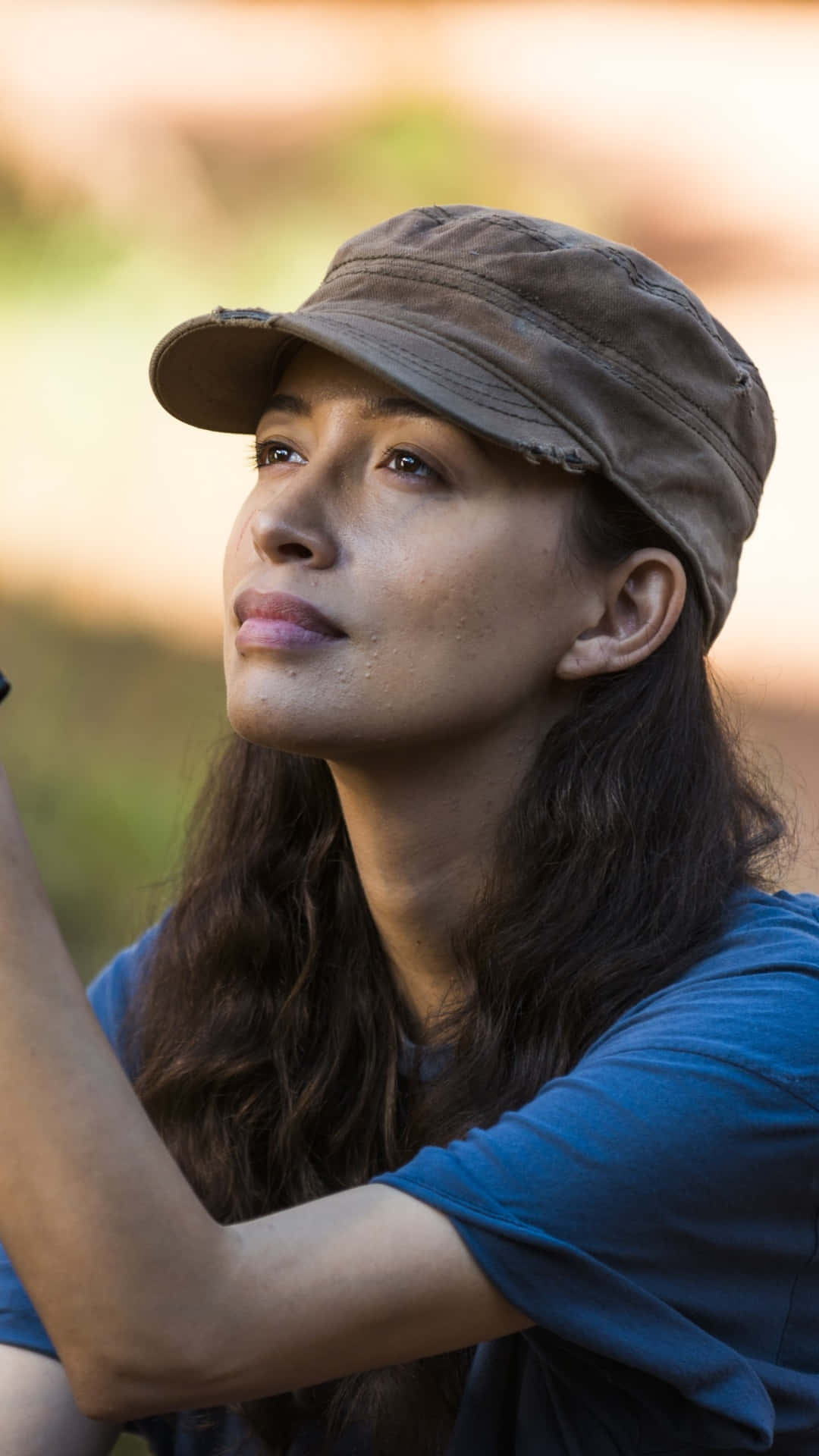 Christian Serratos 桌布 壁紙 — 一位戴著破舊帽子的女性，在柔和模糊的自然背景下，帶著沉思的表情向上凝視。