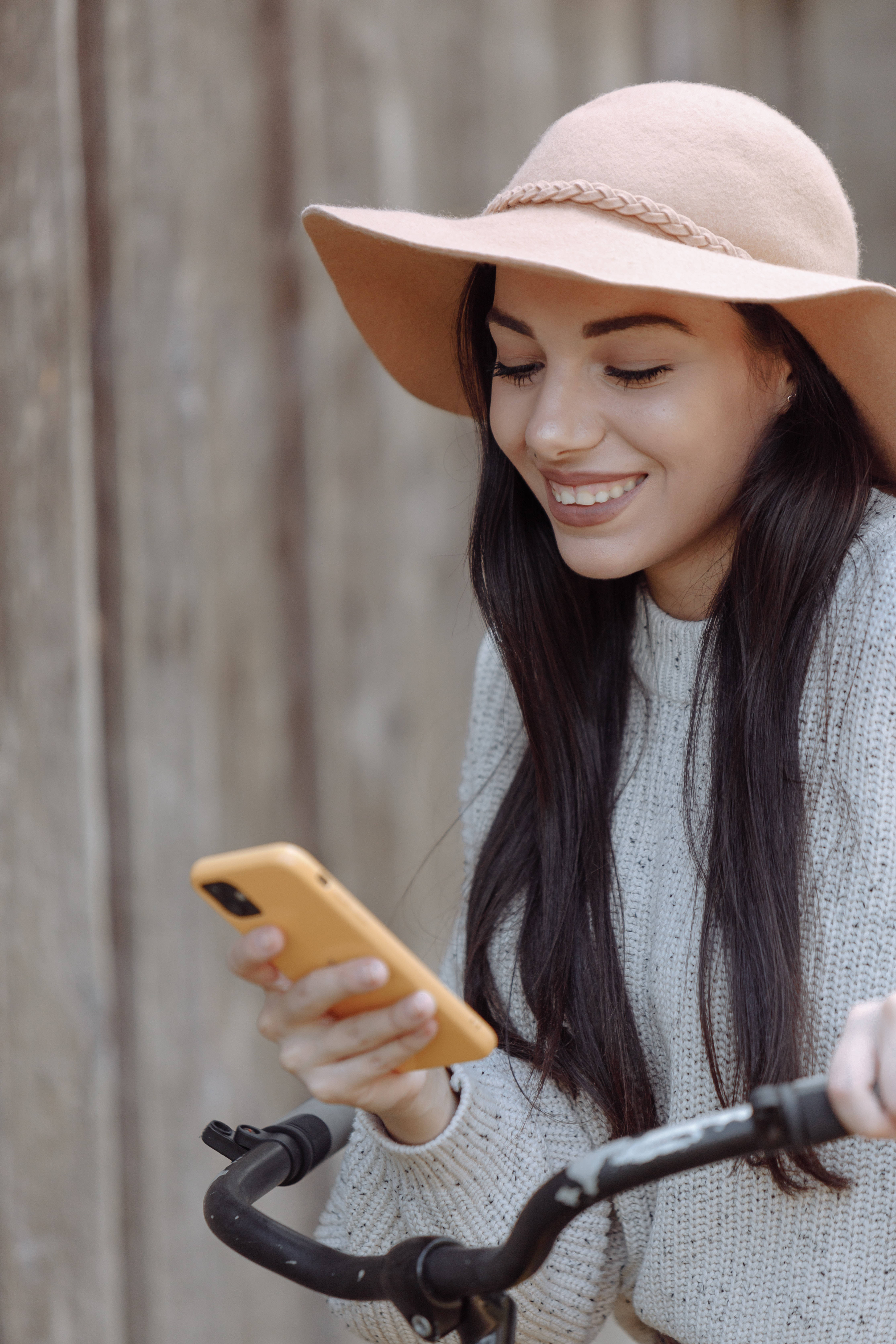酷炫iPhone 桌布 壁紙 — 一位戴著粉紅色帽子和針織毛衣的微笑女士，一邊看著她的黃色手機，一邊握著腳踏車把手靠在木柵欄上。