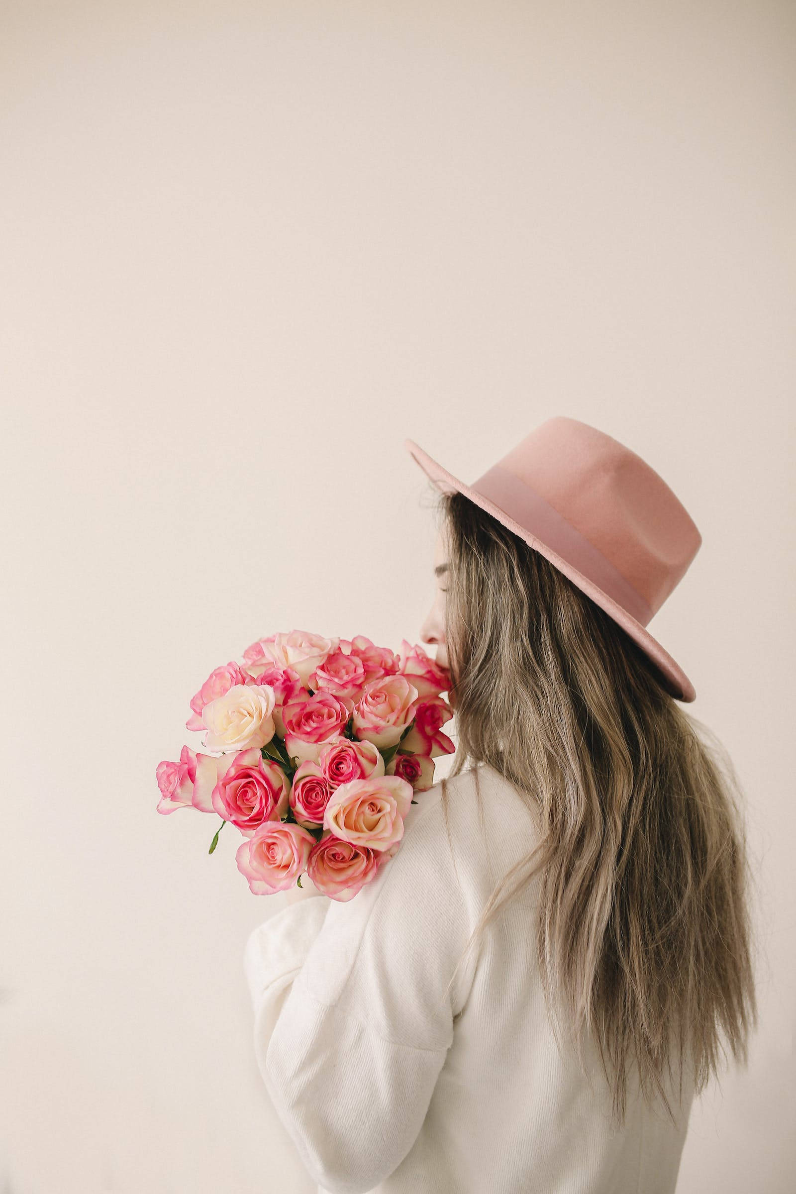 Rose iPhone 桌布 壁紙 — 一位戴著粉紅色帽子的女士，在柔和的米色牆壁前，輕輕聞著一束粉紅和奶油色的玫瑰。