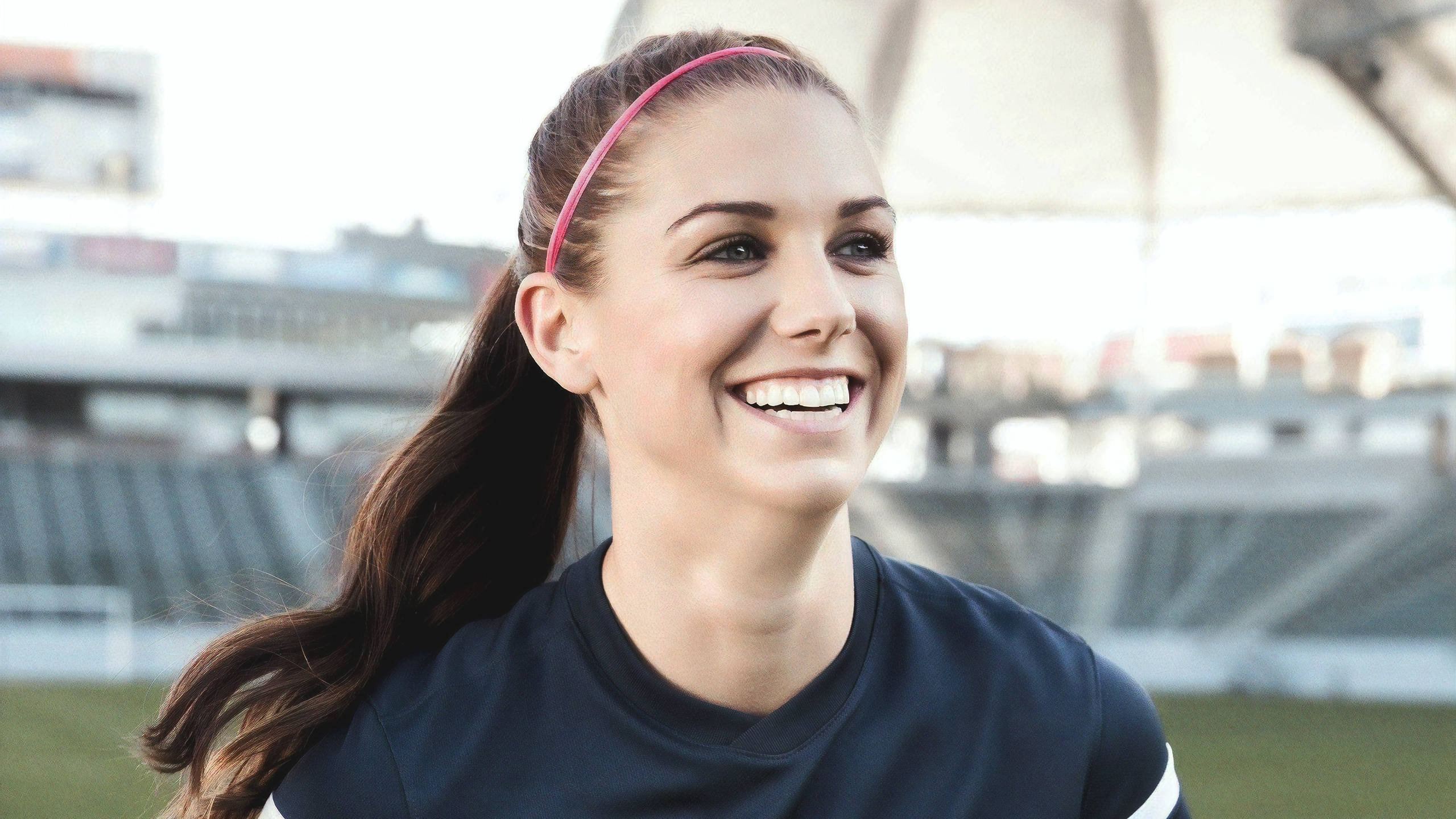 Alex Morgan 桌布 壁紙 — 一位戴著粉紅色頭帶的快樂年輕女性，在陽光明媚的體育場穿著深色運動衫，燦爛地微笑。