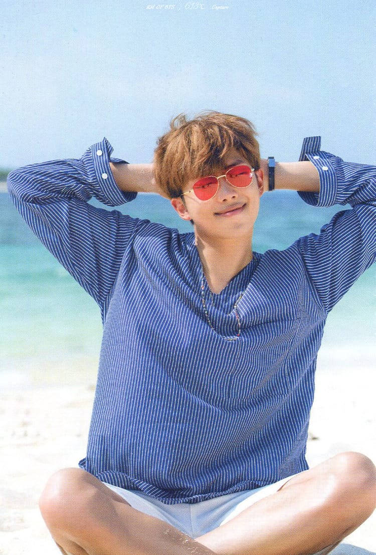 BTS 桌布 壁紙 — 一位戴著紅色太陽眼鏡、身穿藍色條紋襯衫的年輕男子，在陽光普照的海灘上盤腿而坐，身後是碧綠的海水。