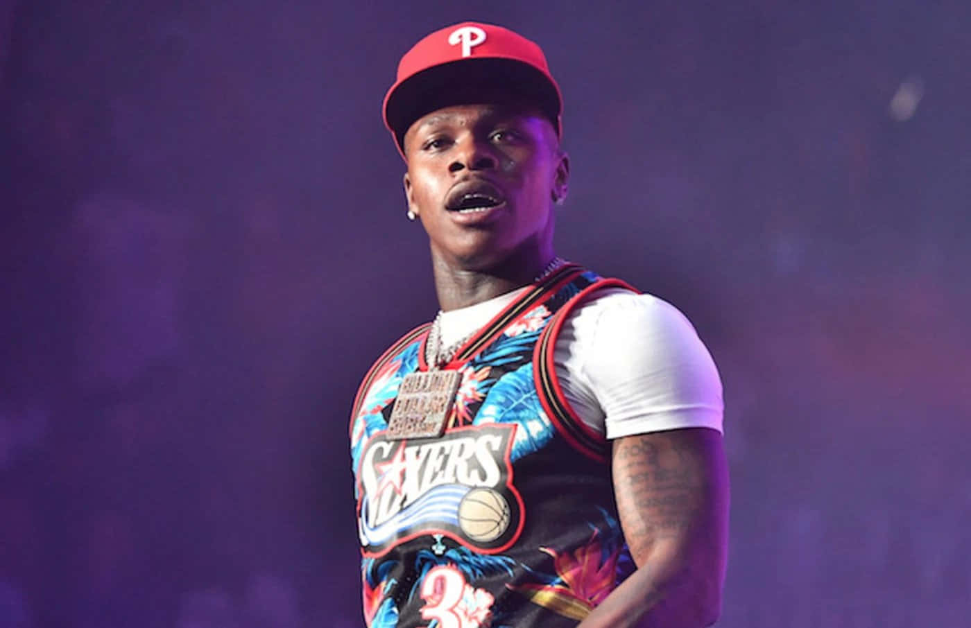 Dababy 桌布 壁紙 — 一位戴著紅色帽子、身穿彩色籃球球衣的男性表演者在紫色舞台燈光下唱歌。