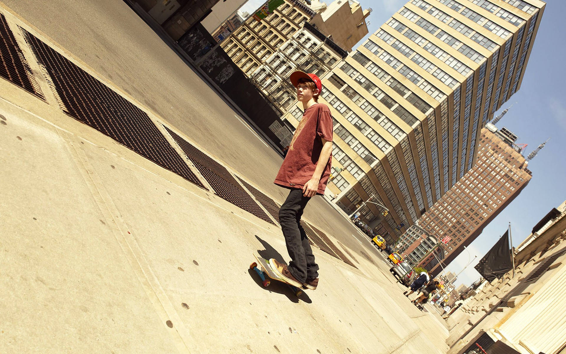 Skater Boy 桌布 壁紙 — 一位戴著紅色帽子的年輕滑板手，在高樓大廈環繞的陽光普照城市廣場上滑行。