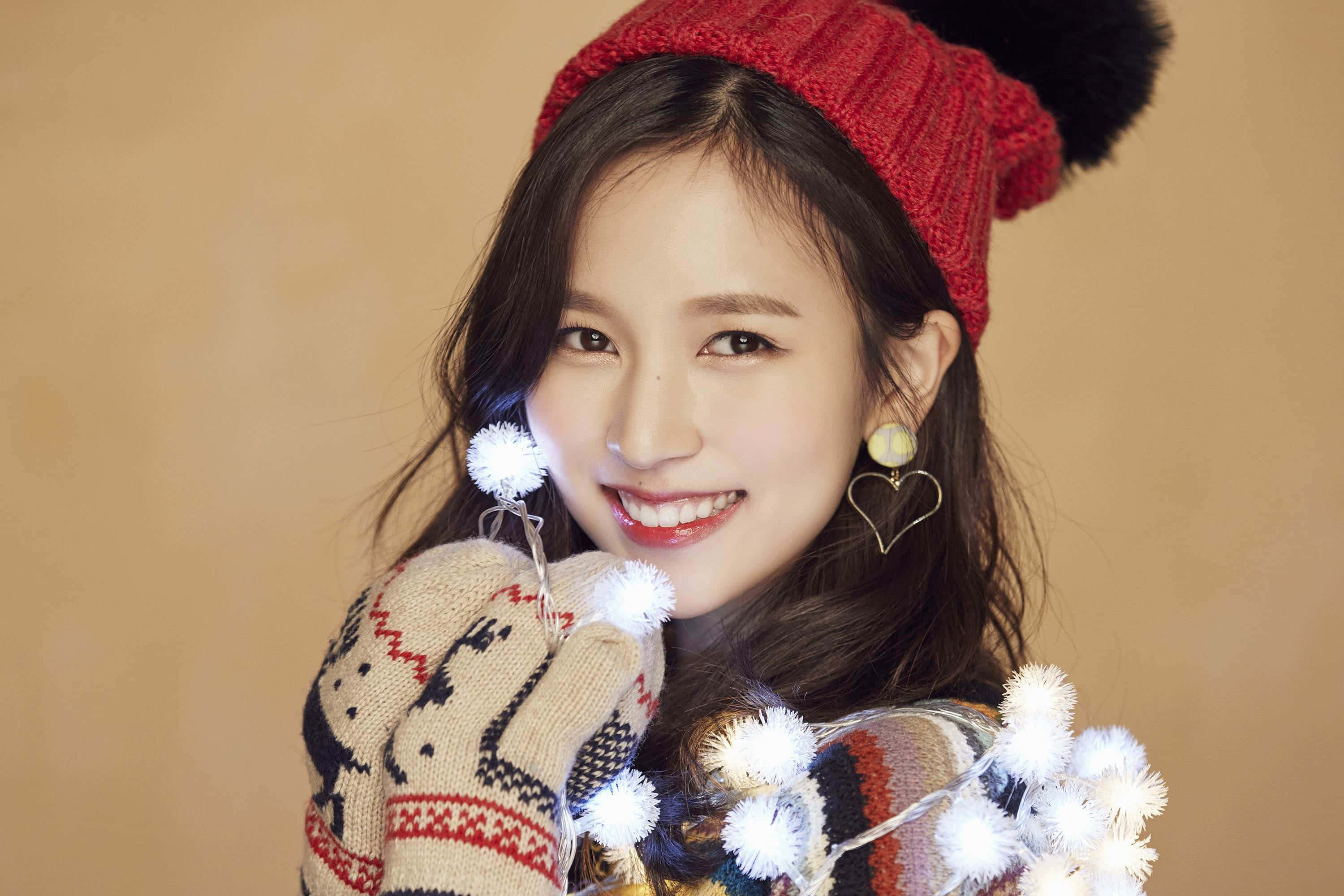 twice 桌布 壁紙 — 一位戴著紅色針織毛帽和圖案手套的開朗年輕女子，微笑著拿著一串白色絨球燈。