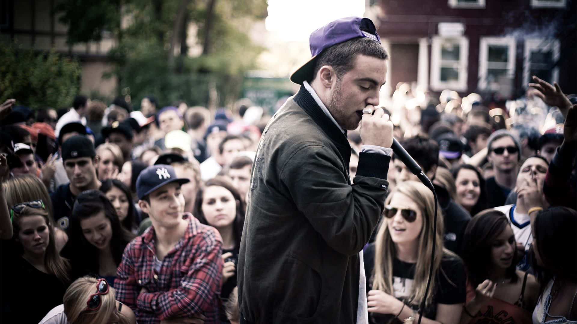 Mac Miller 桌布 壁紙 — 一位戴著紫色帽子的饒舌歌手在戶外對著麥克風表演，面前是充滿活力的觀眾。