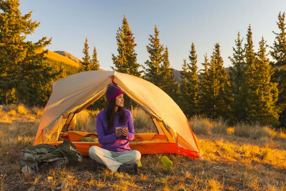 Fall Camping 桌布 壁紙 — 一位戴著紫色毛帽的女性盤腿坐在橙色帳篷外，啜飲咖啡，金色夕陽灑在松樹和山巒上。