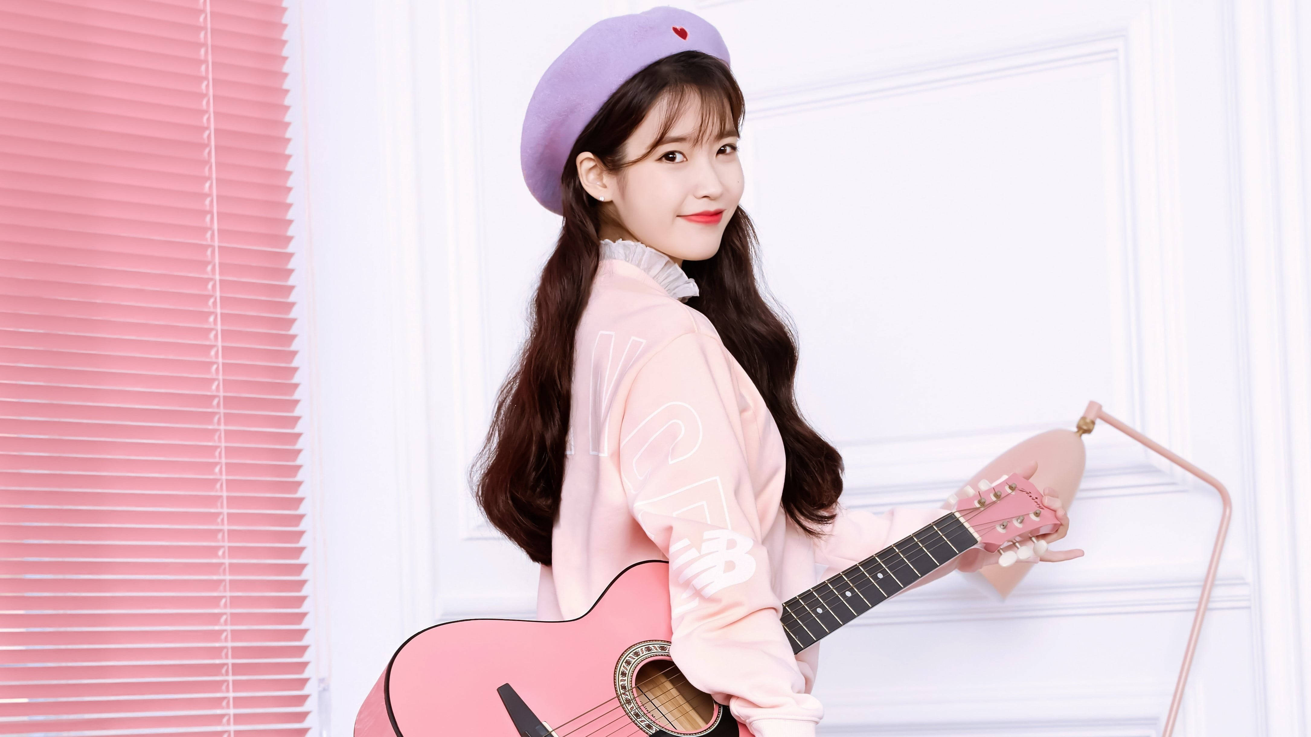 Kpop Aesthetic 桌布 壁紙 — 一位戴著紫色貝雷帽、身穿粉紅色外套的年輕女子，在粉紅色百葉窗和白色門旁拿著一把粉紅色木吉他。