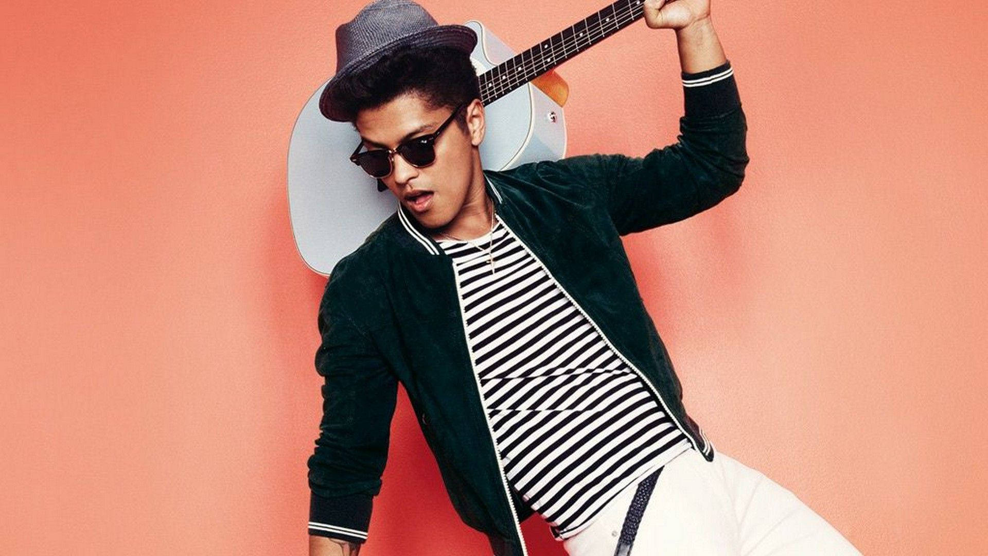 Bruno Mars 桌布 壁紙 — 一位戴著紳士帽和太陽眼鏡的時尚男士，在珊瑚粉紅背景前自信地擺姿勢，手持木吉他。