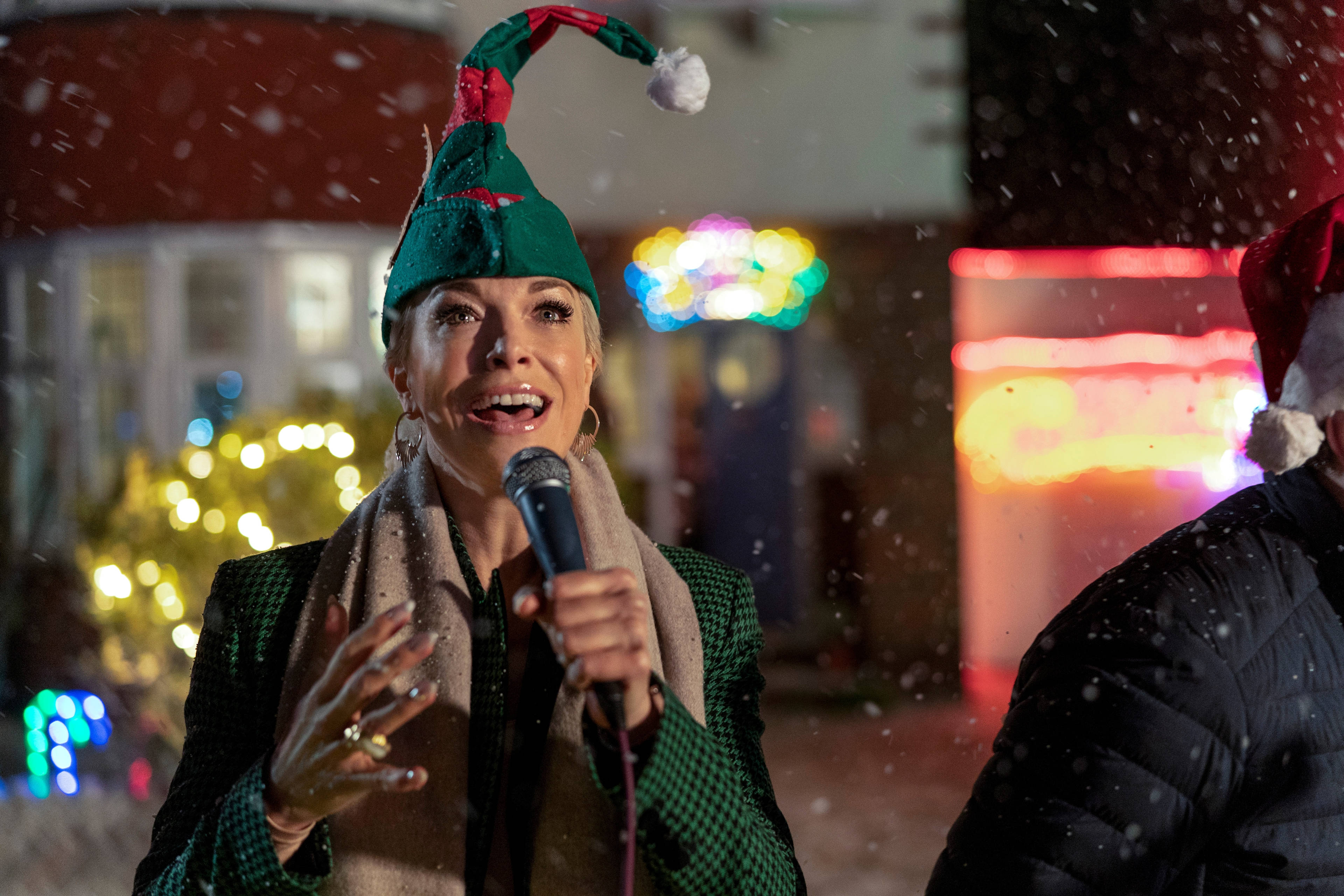 Ted Lasso 桌布 壁紙 — 一位戴著綠色精靈帽子的女士在夜晚雪花飄落、節日燈光模糊的背景下，對著麥克風歡快地唱歌。