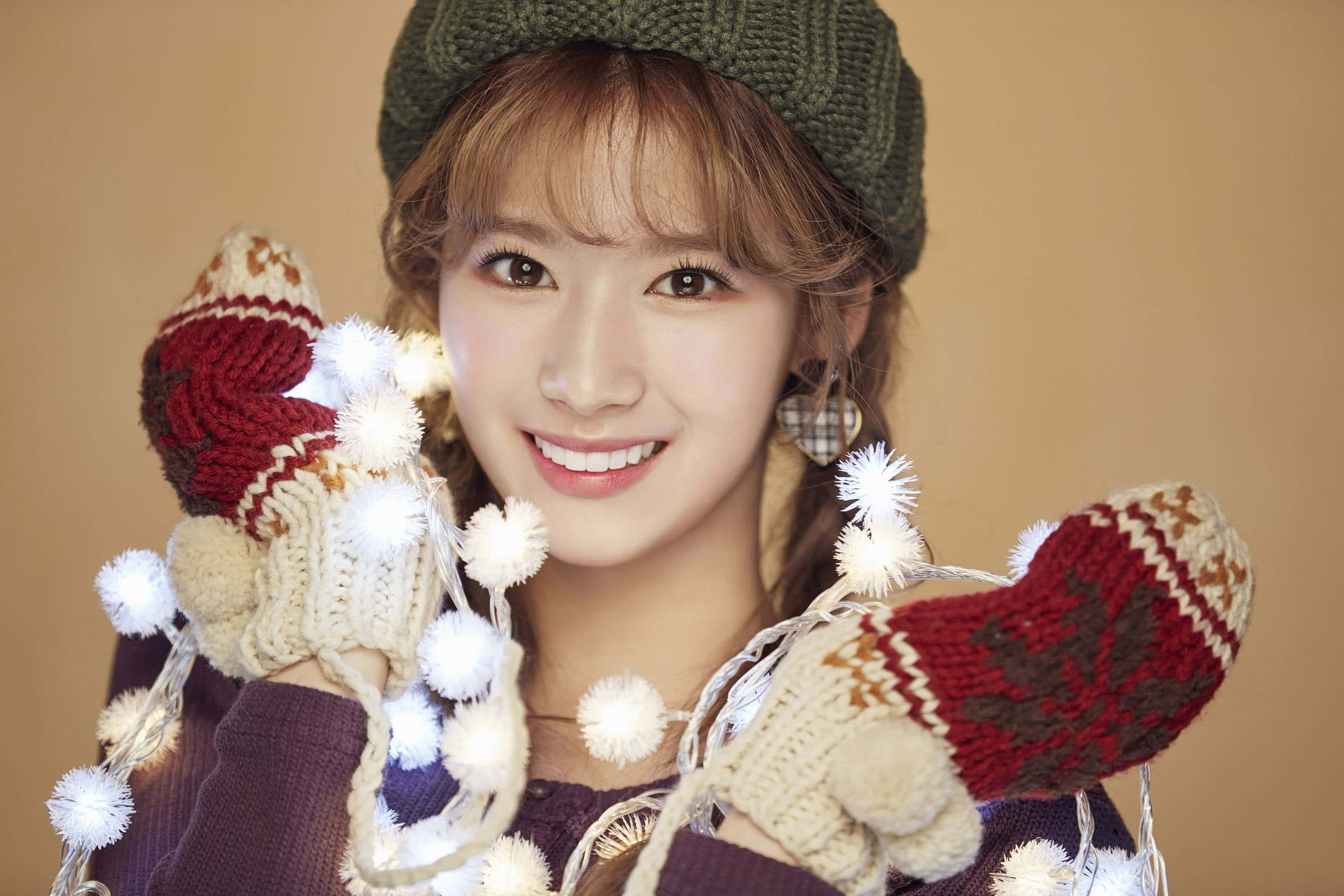 twice 桌布 壁紙 — 一位戴著綠色針織帽和節慶手套的開朗年輕女性，手持發光的燈串，溫暖地微笑。