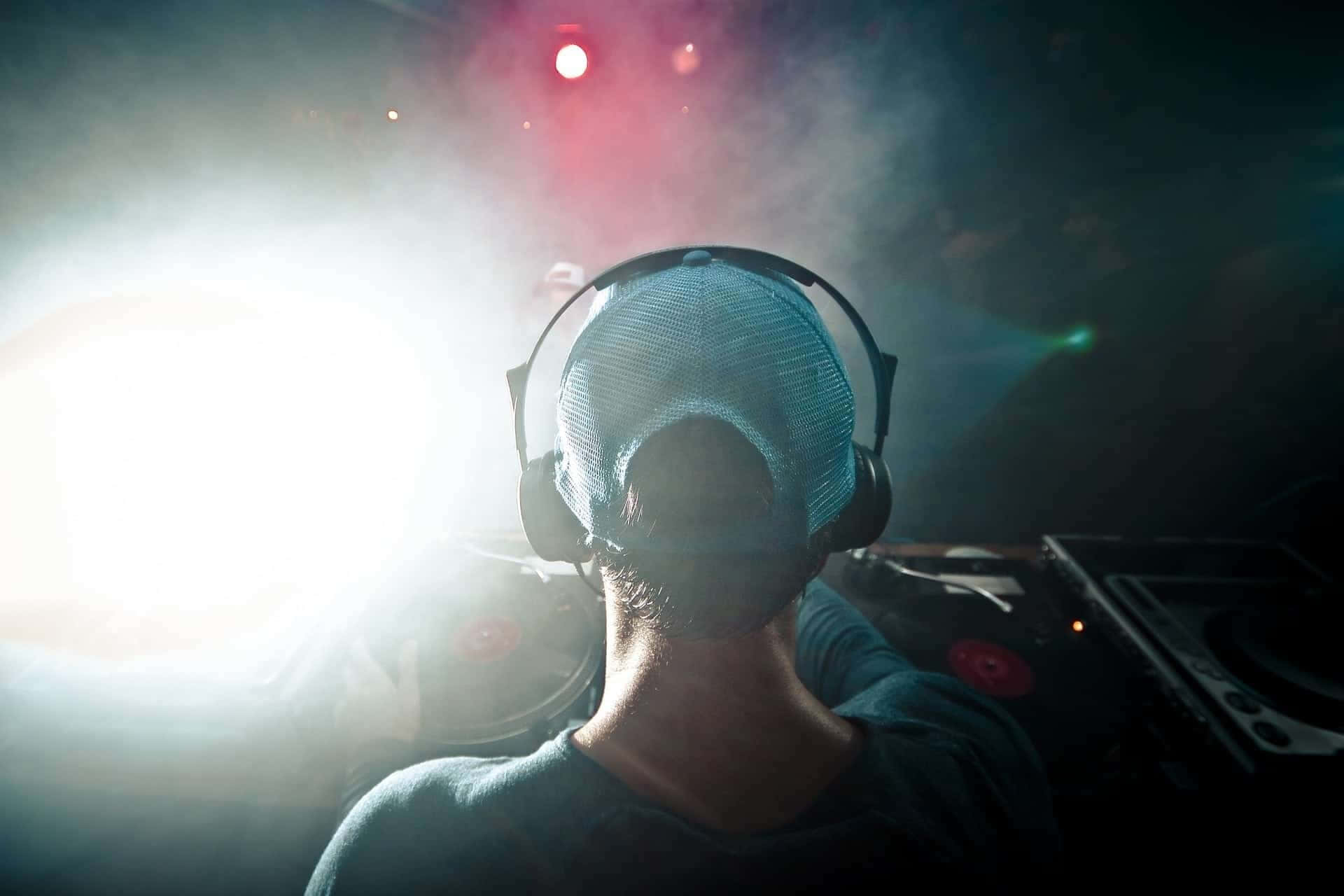 concert 桌布 壁紙 — 一位戴著網帽和耳機的DJ在黑暗的俱樂部中面對著明亮的舞台燈光，伴隨著紅色聚光燈和煙霧。