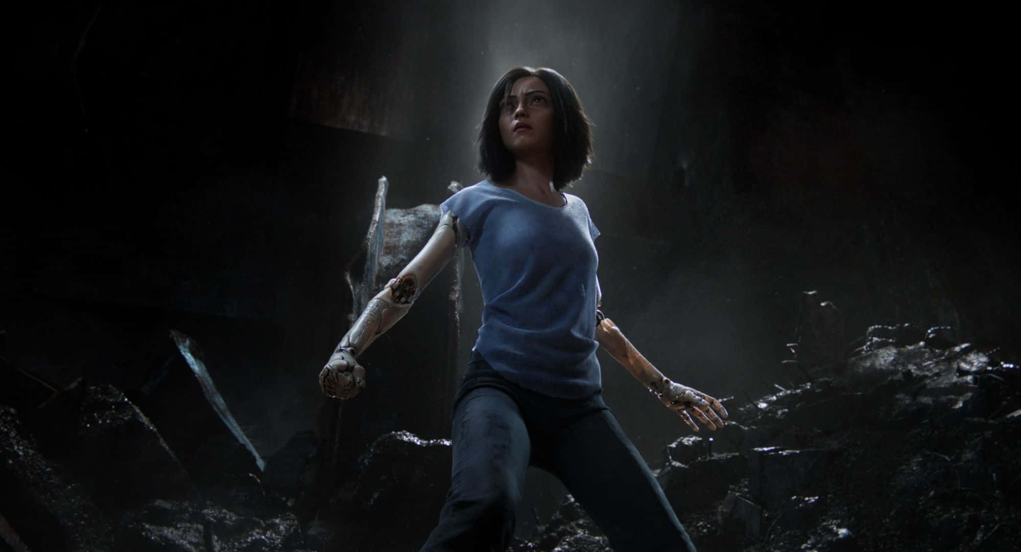 Alita Battle Angel 桌布 壁紙 — 一位戴著義肢手臂的女性站在黑暗的廢墟環境中，被戲劇性的光束照亮。