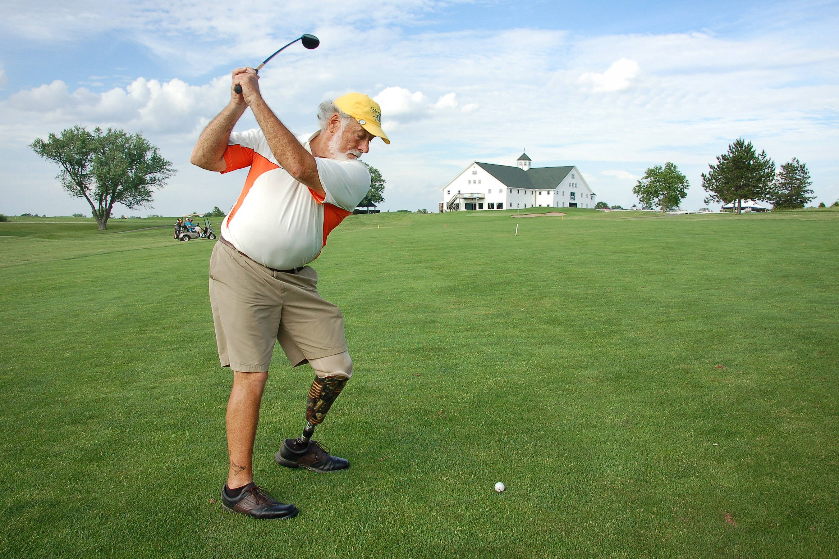 4K Golf 桌布 壁紙 — 一位戴著義肢的長者，在陽光明媚的綠色球場上揮動高爾夫球桿，遠處有一座白色農舍。