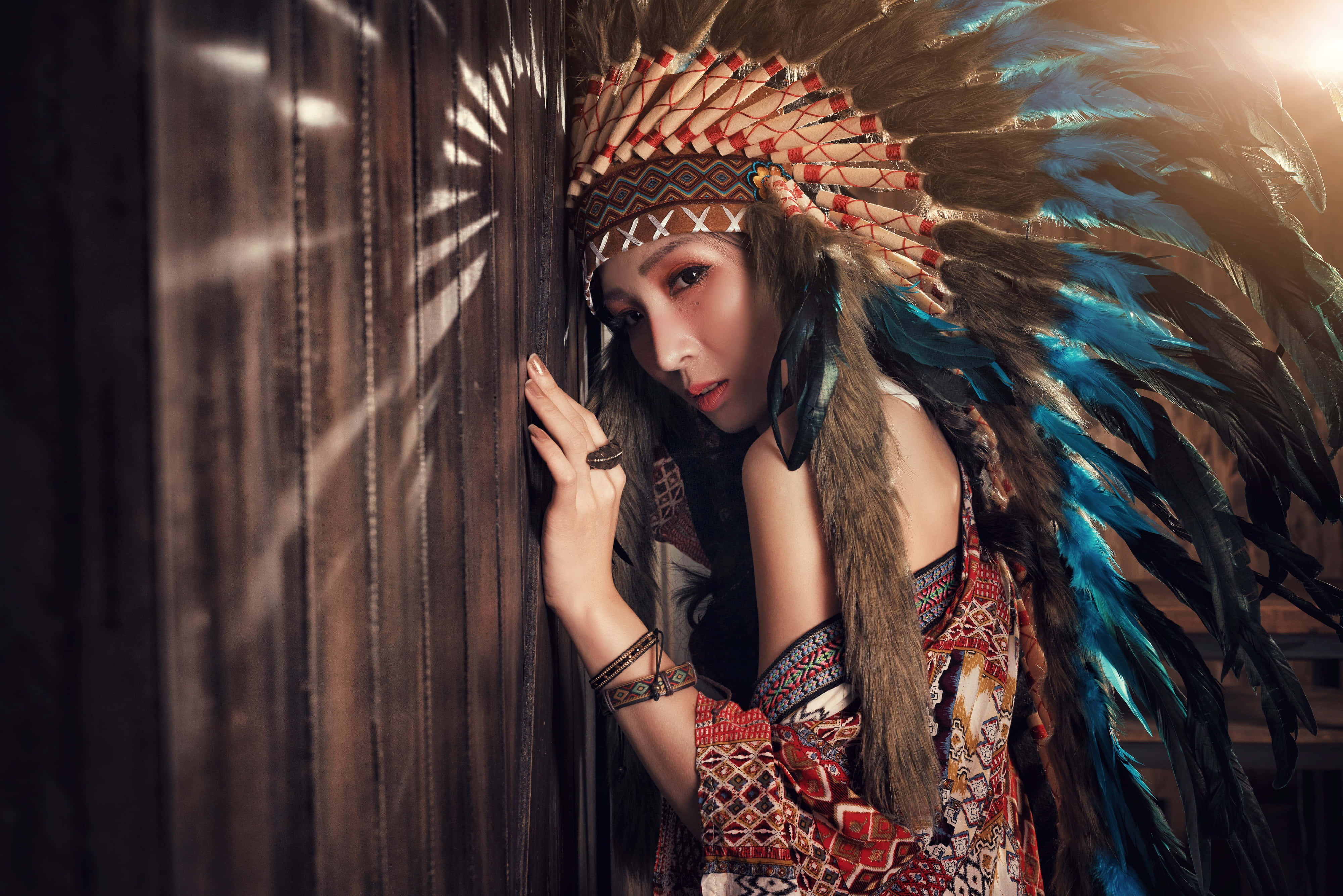 Woman 桌布 壁紙 — 一位戴著羽毛頭飾的女人靠在一扇木門上，溫暖的陽光從她身後的縫隙中照射進來。