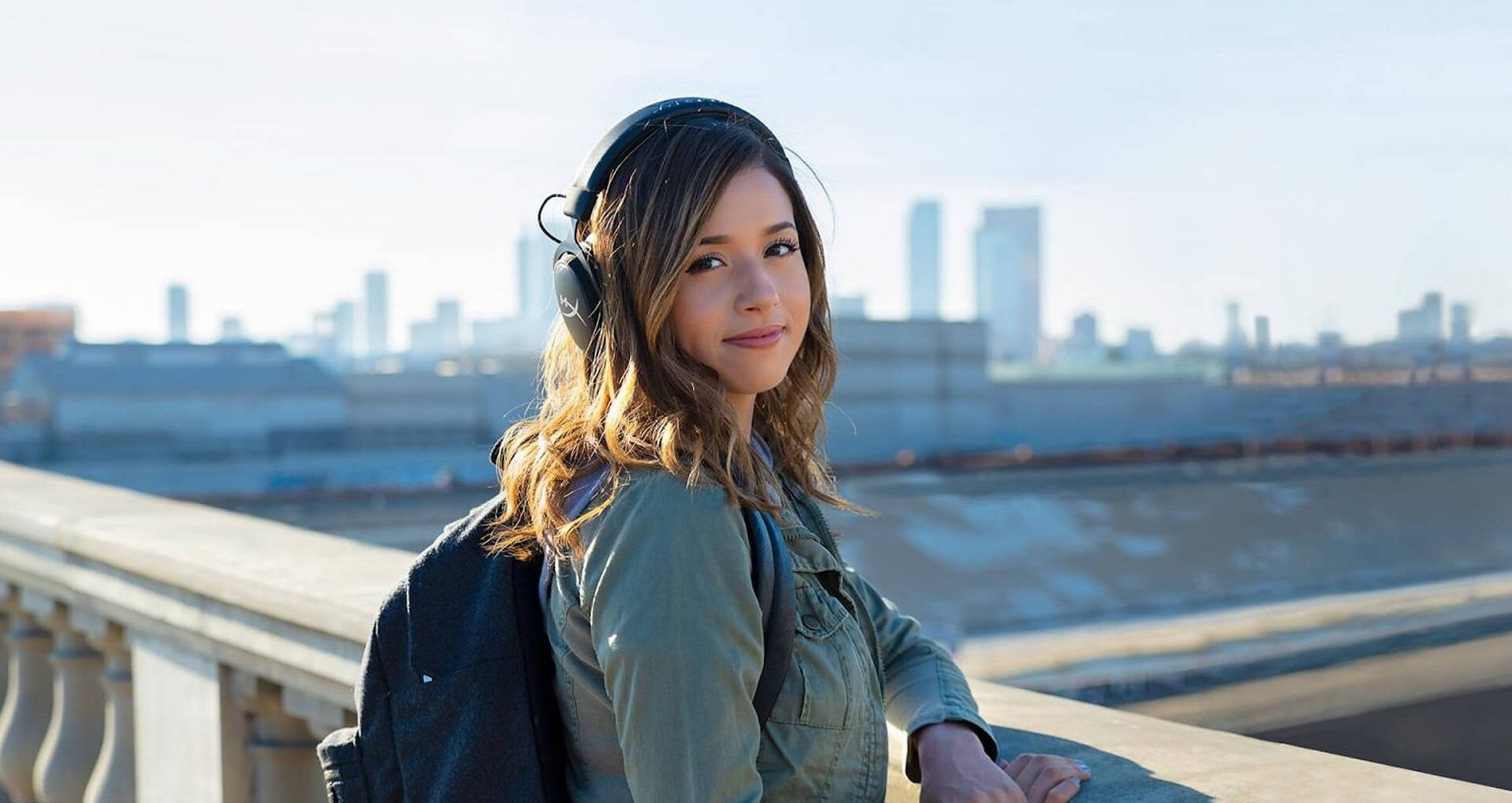 pokimane 桌布 壁紙 — 一位戴著耳機的年輕女子微笑著靠在欄杆上，俯瞰著明亮的城市天際線。