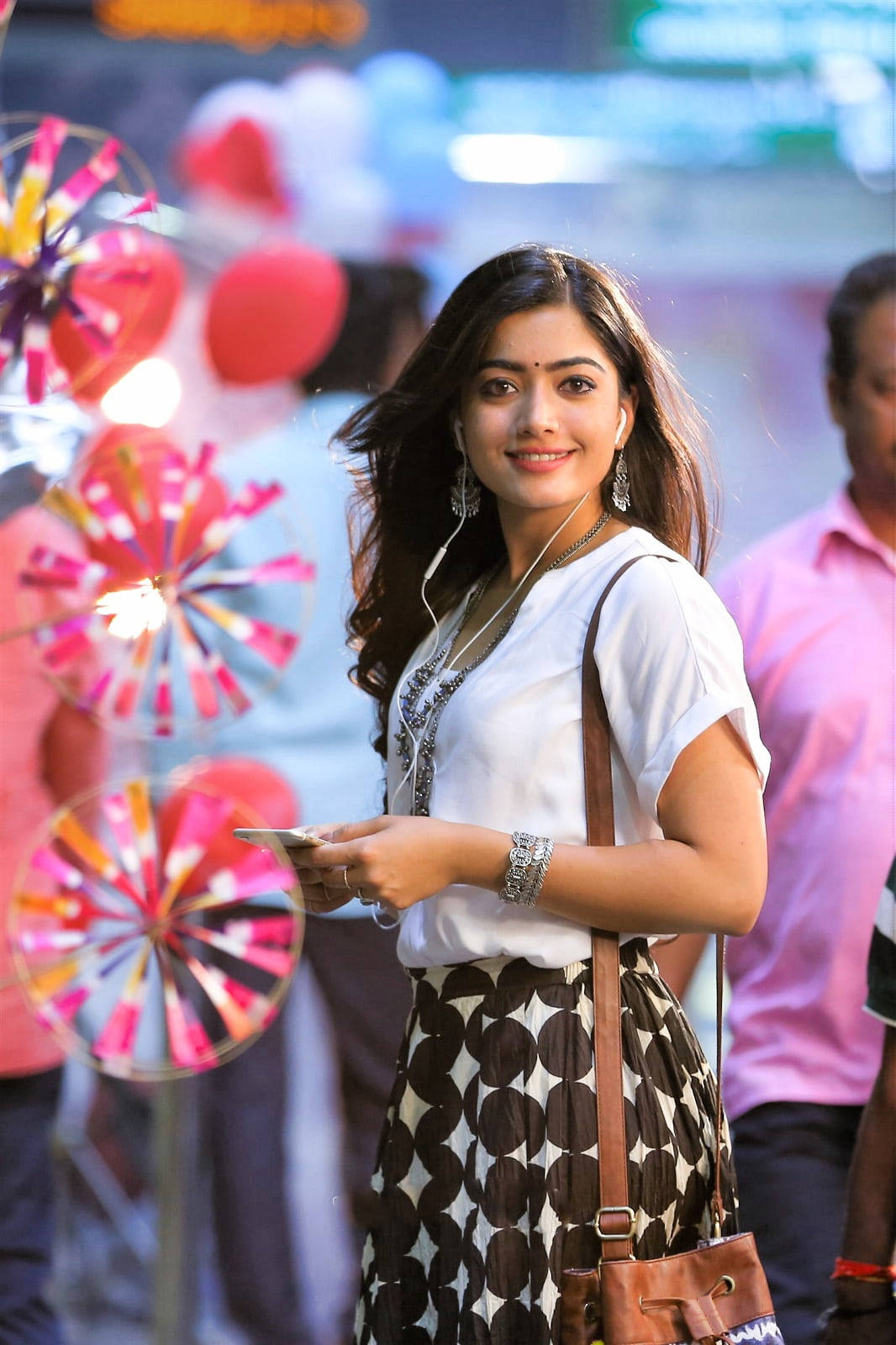 telugu 桌布 壁紙 — 一位戴著耳機的年輕微笑女子走過一條裝飾著彩色旋轉風車和模糊行人的繁忙街道。