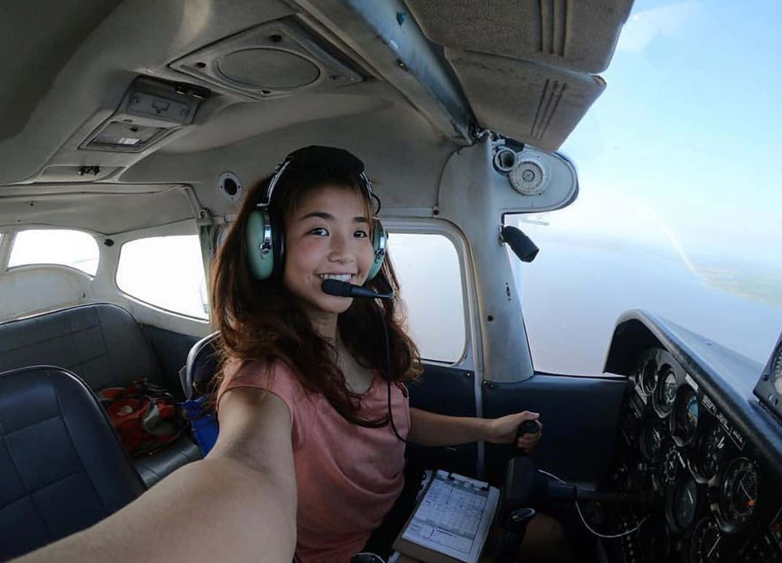 Plane 桌布 壁紙 — 一位戴著耳機的年輕微笑女性駕駛小型飛機，透過駕駛艙窗戶可以看到晴朗的天空和雲朵。