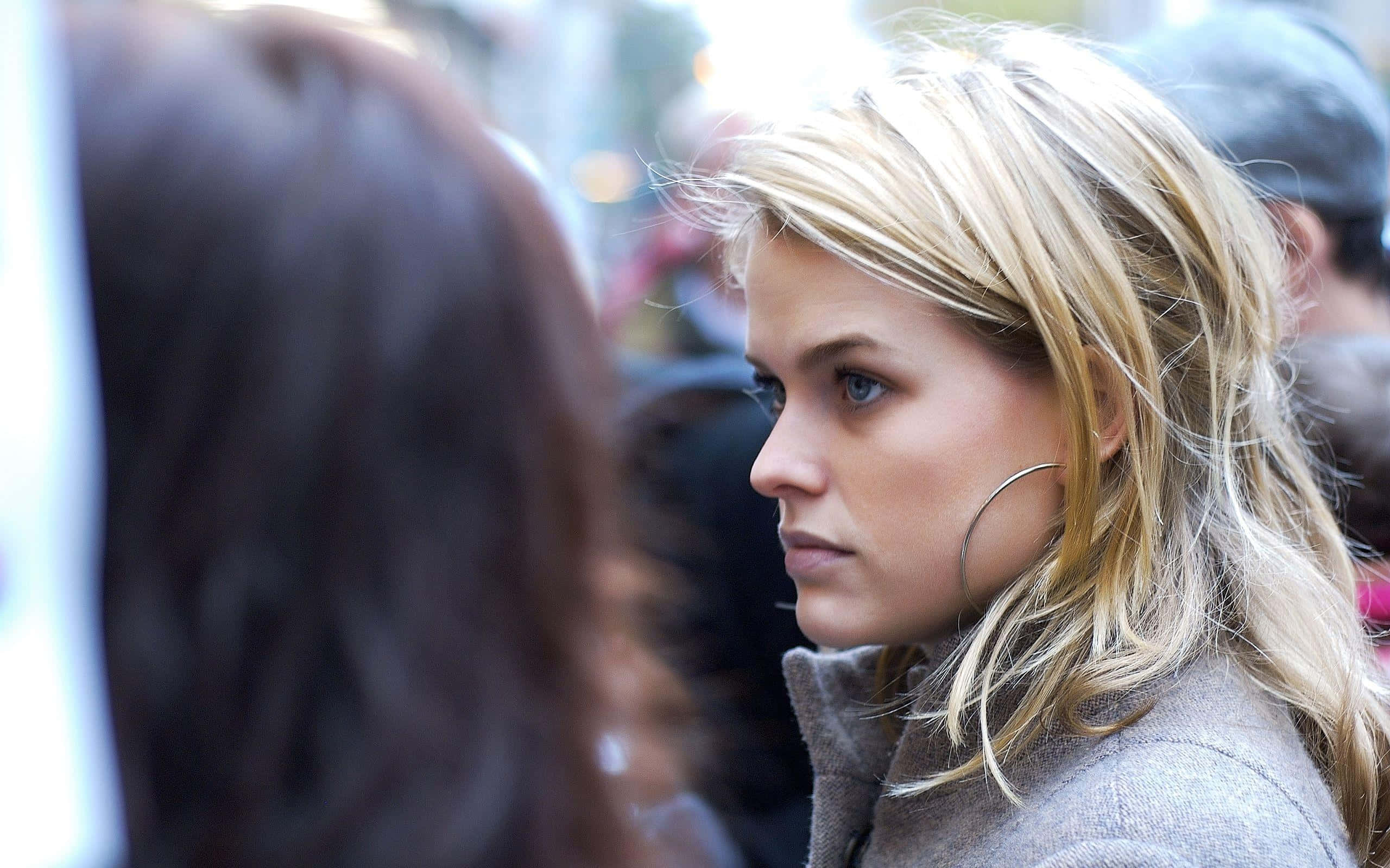 alice eve 桌布 壁紙 — 一位戴著耳環的金髮女子穿過模糊的城市人群，專注地凝視著某人。