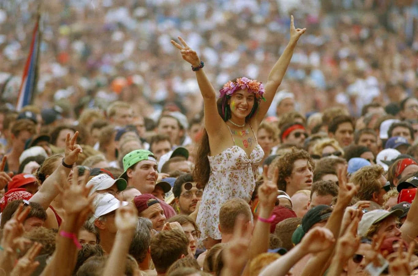 woodstock 桌布 壁紙 — 一位戴著花冠、比出和平手勢的快樂女性，在明亮的日光下從一片歡呼的演唱會人群中閃耀。