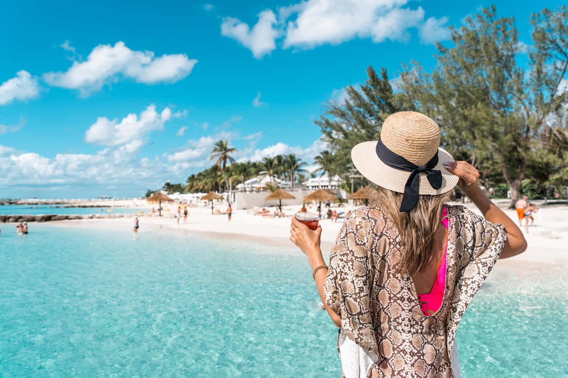Bahamas Island 桌布 壁紙 — 一位戴著草帽的女士在明亮的藍天下，在棕櫚樹和白色沙灘的藍綠色海灘上啜飲飲料。