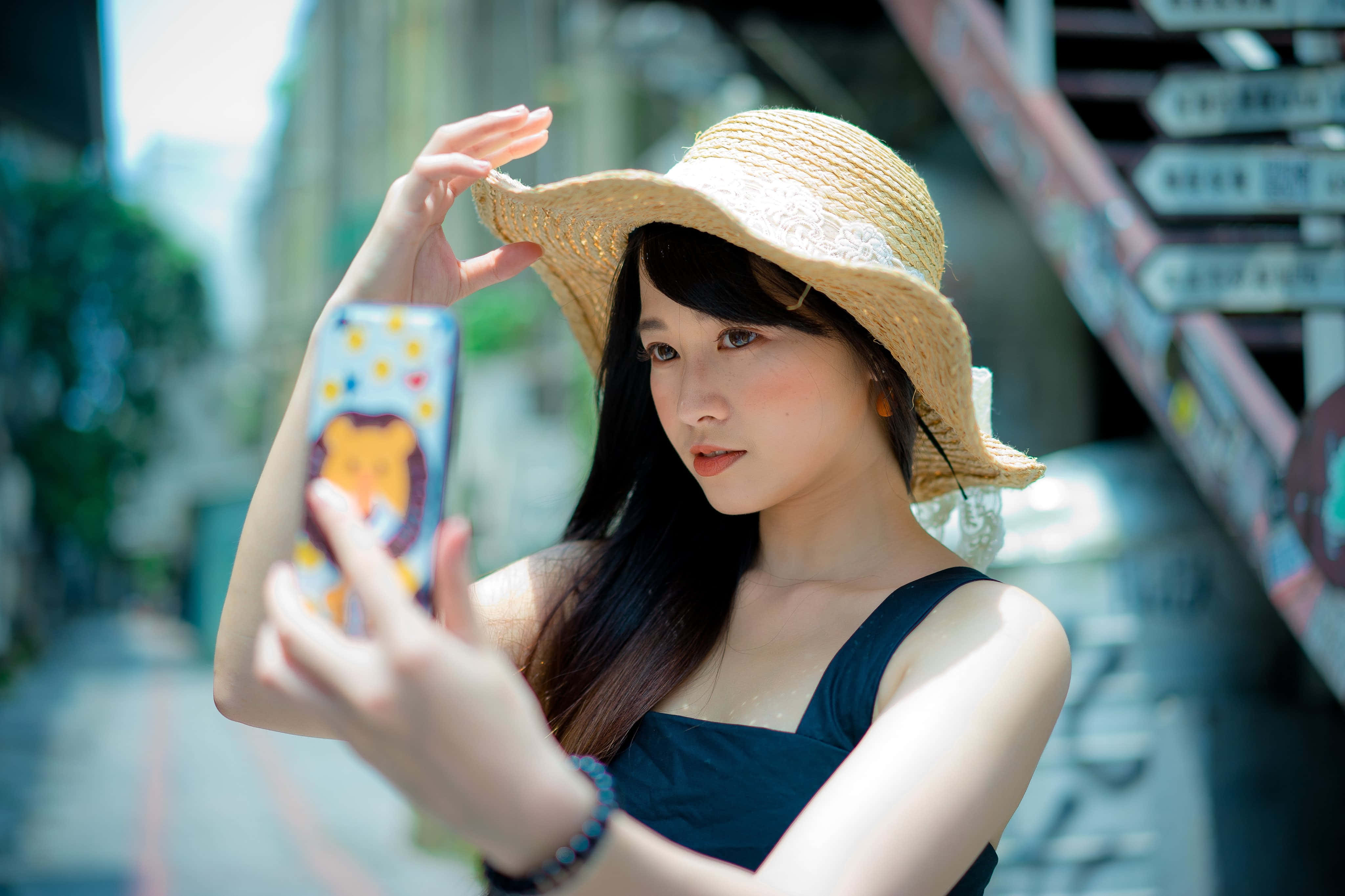 Summer Fashion 桌布 壁紙 — 一位戴著草帽的年輕女性在陽光明媚的城市街道上，用色彩繽紛的手機殼自拍。