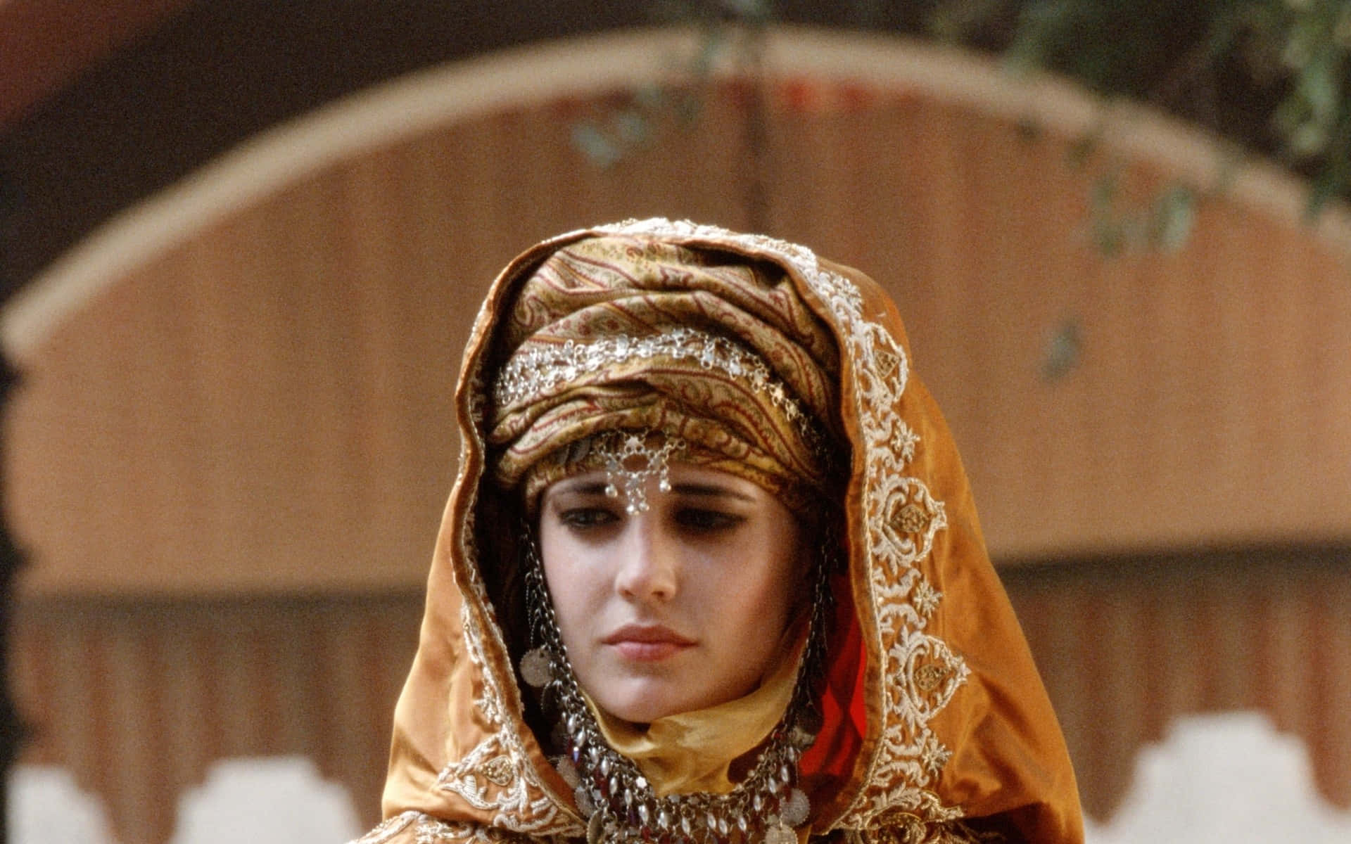 eva green 桌布 壁紙 — 一位戴著華麗金色頭飾和珠寶的女性，在木製拱形背景下，表情莊嚴地向下凝視。