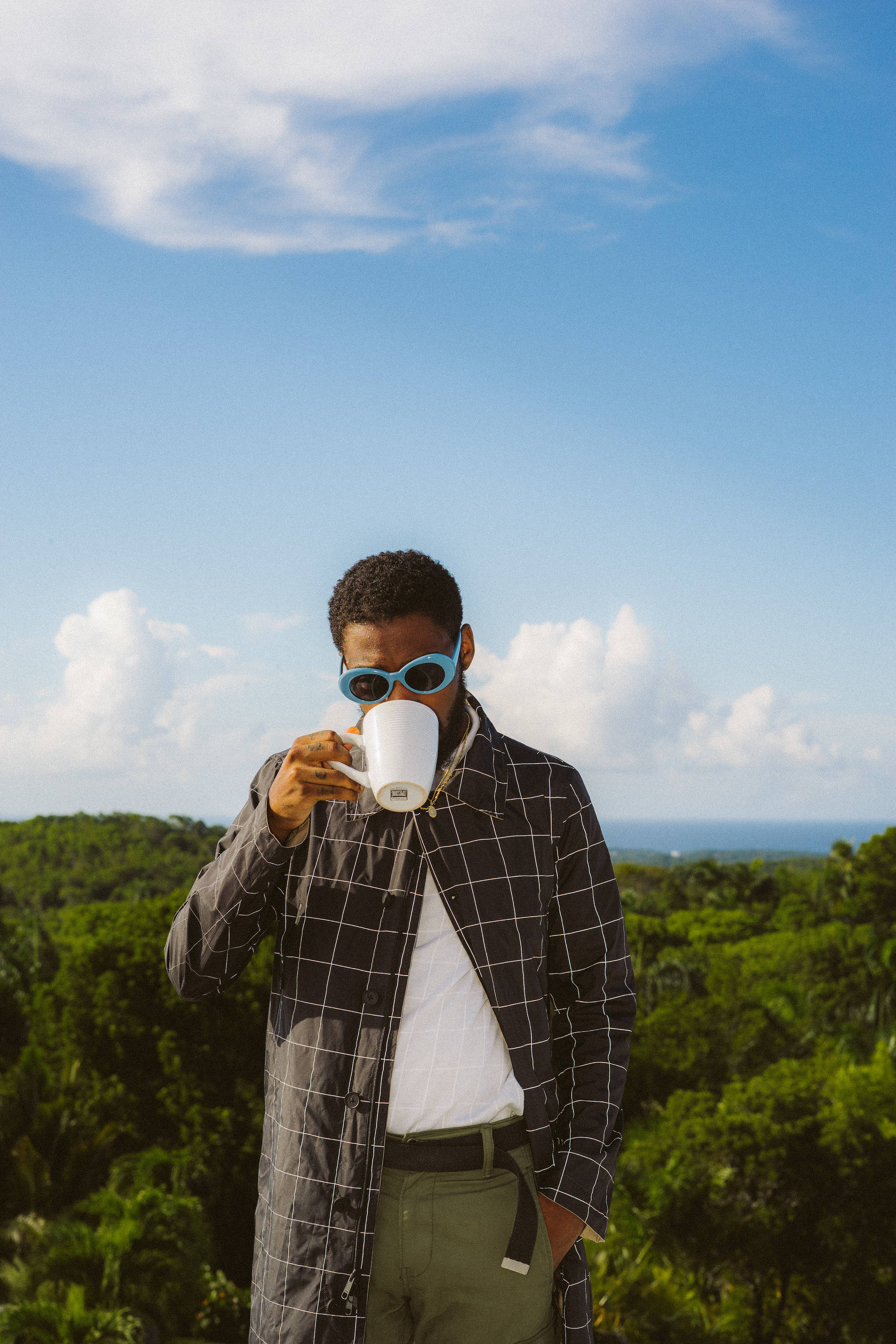 Brent Faiyaz 桌布 壁紙 — 一位戴著藍色太陽眼鏡的時尚男士，在翠綠的山丘上俯瞰海洋，在明亮的藍天下啜飲咖啡。