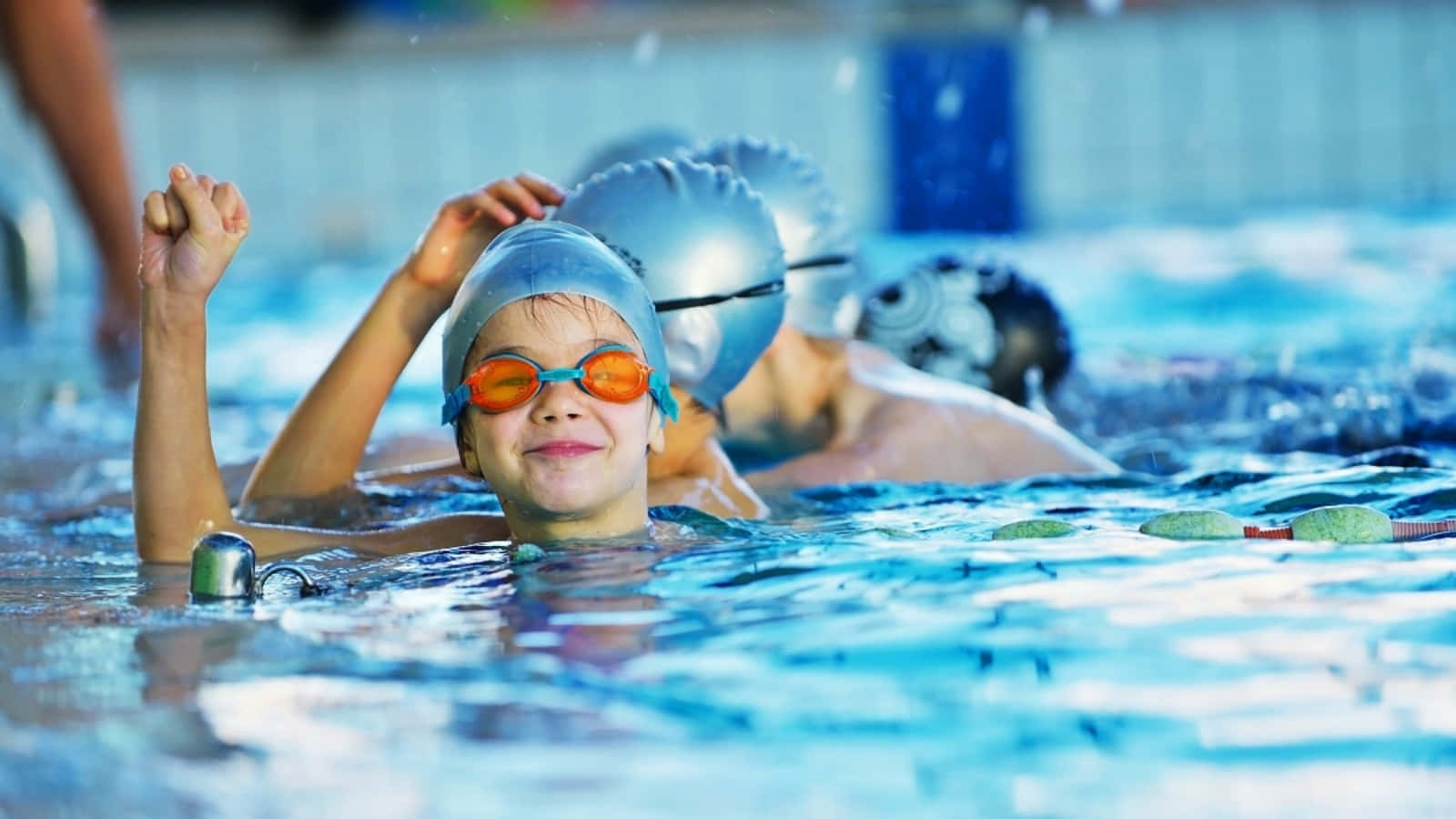Swimming 桌布 壁紙 — 一位戴著藍色泳帽和橙色泳鏡的年輕泳者，在泳池中高舉拳頭，露出勝利的笑容。