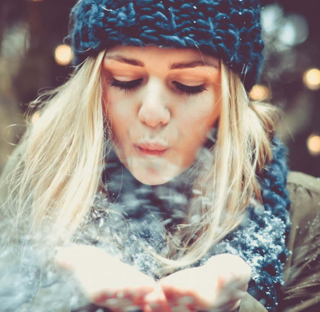 冬天 桌布 壁紙 — 一位戴著藍色針織帽的女性，從她捧著的雙手中吹出雪花，背景是柔和模糊的冬日景色，帶有溫暖的散景燈光。