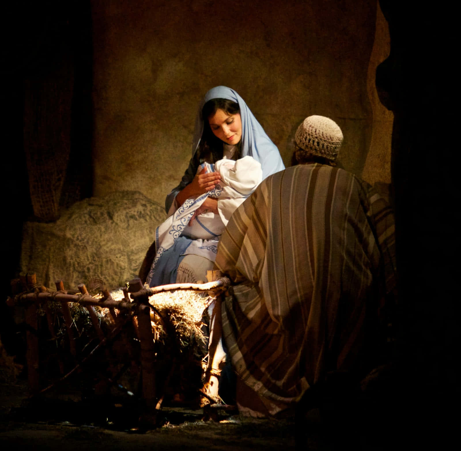 Nativity 桌布 壁紙 — 一位戴著藍色面紗的女子溫柔地抱著一個包裹著的嬰兒，而一位身穿條紋長袍的男子跪在昏暗馬廄裡一個簡陋的馬槽旁。