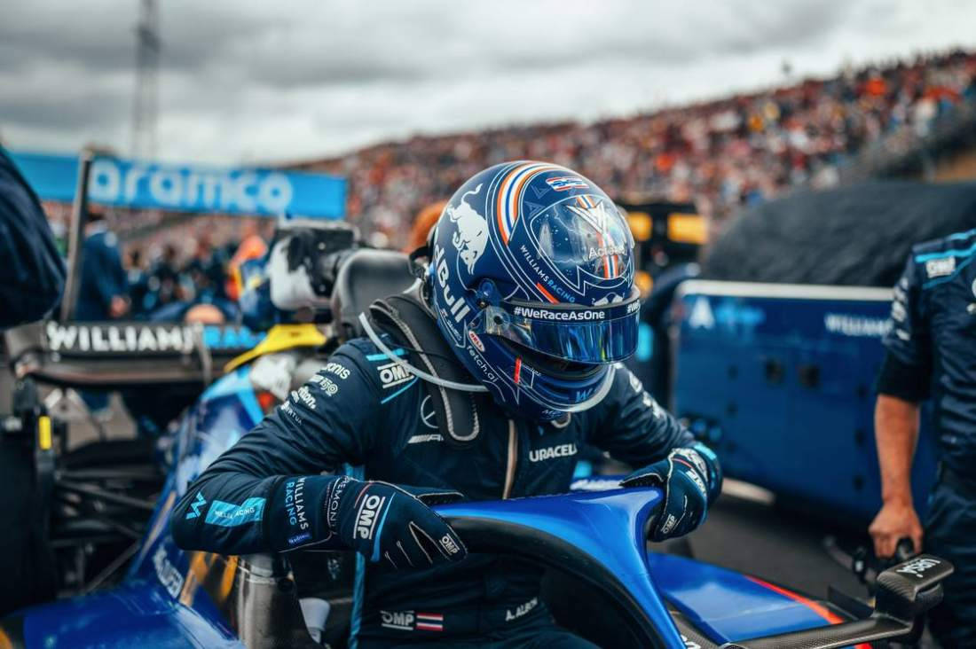 F1 桌布 壁紙 — 一位戴著藍色頭盔和穿著藍色賽車服的F1車手，在陰天擁擠的維修區車道上靠在他的藍色賽車上。