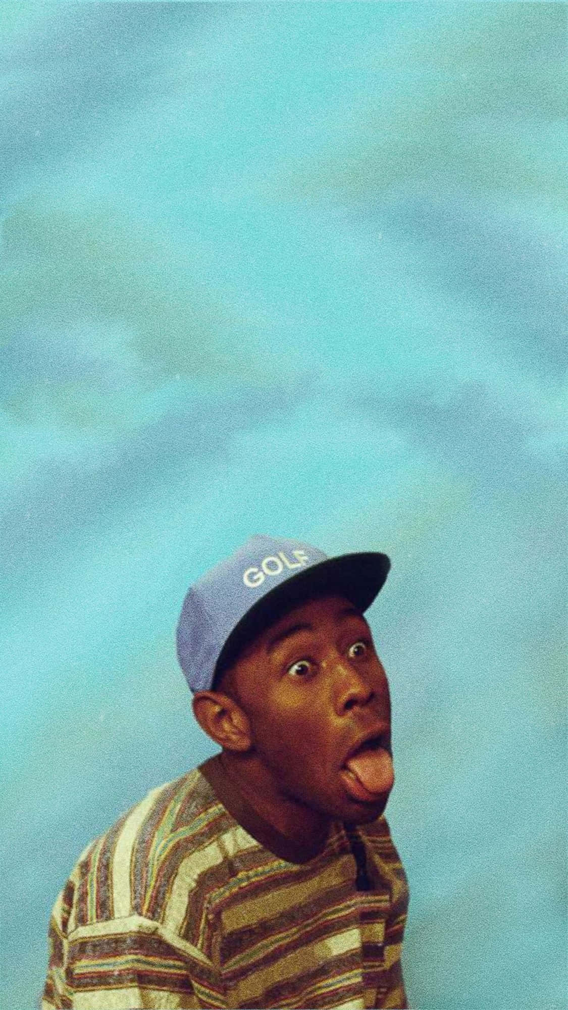 Tyler The Creator 4K 桌布 壁紙 — 一位戴著藍色鴨舌帽、身穿條紋襯衫的年輕男子，在柔和的藍天背景下伸出舌頭。
