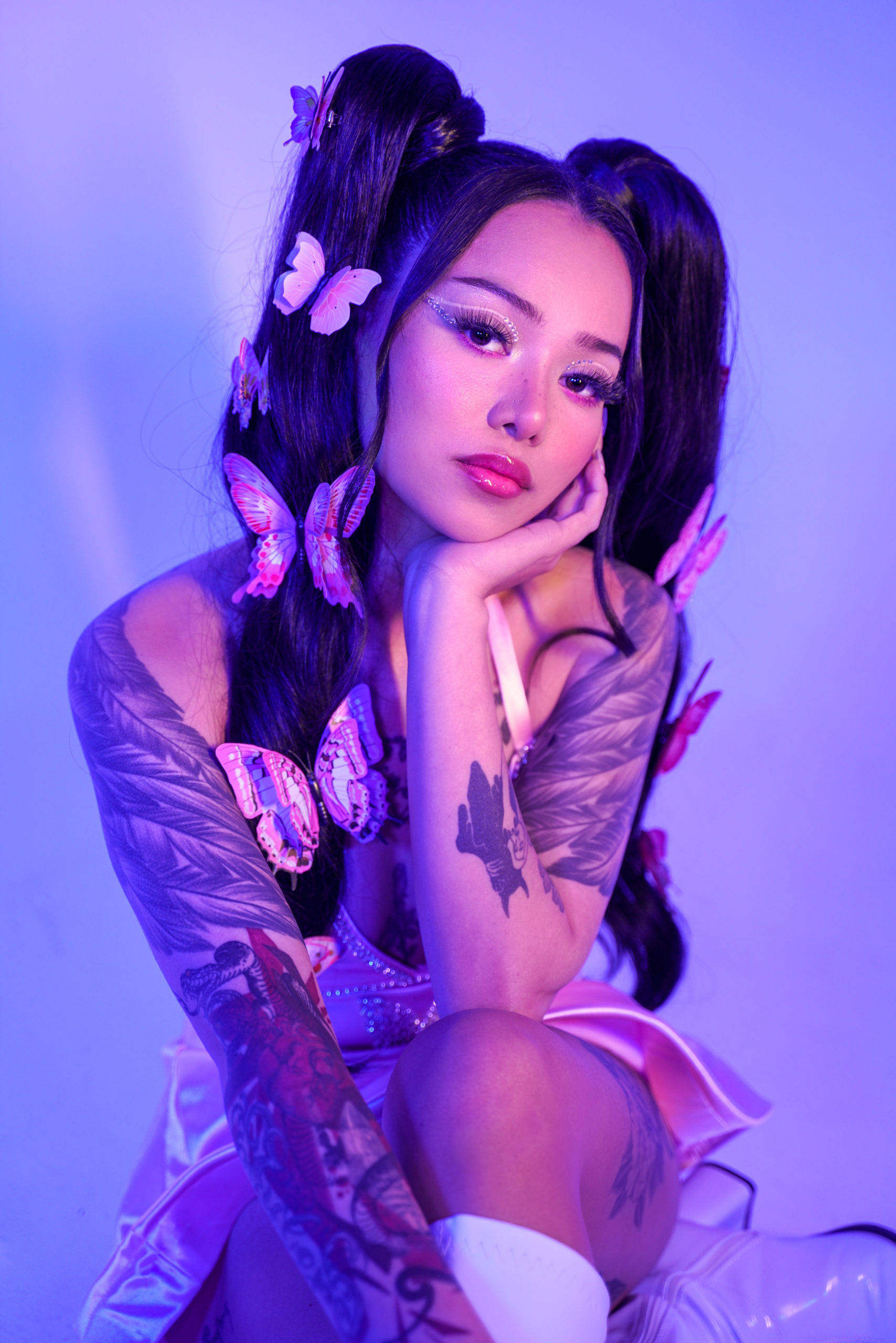 Bella Poarch 桌布 壁紙 — 一位戴著蝴蝶髮夾和刺青的女性在工作室的紫色燈光下擺姿勢。