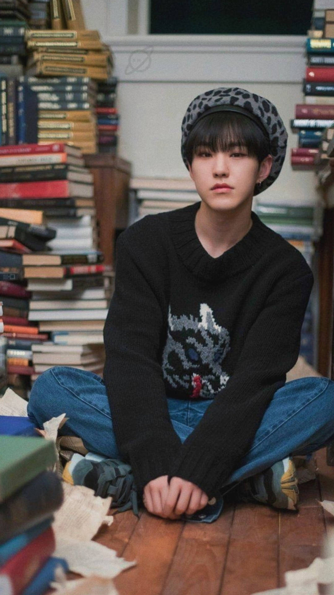 Hoshi 桌布 壁紙 — 一位戴著豹紋貝雷帽和黑色毛衣的年輕人，在舒適的圖書館中盤腿坐在高聳的書堆之間。