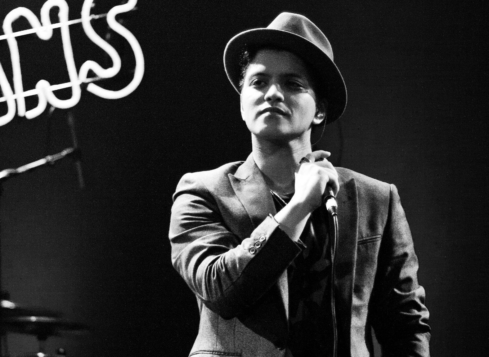 Bruno Mars 桌布 壁紙 — 一位戴著費多拉帽和西裝的歌手在霓虹燈下的舞台上拿著麥克風的黑白肖像。