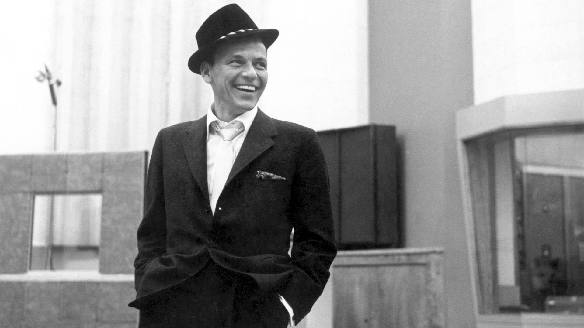 Sinatra 桌布 壁紙 — 一位戴著軟呢帽、身穿西裝的微笑男子，自信地站在現代主義建築外牆前，黑白照片。